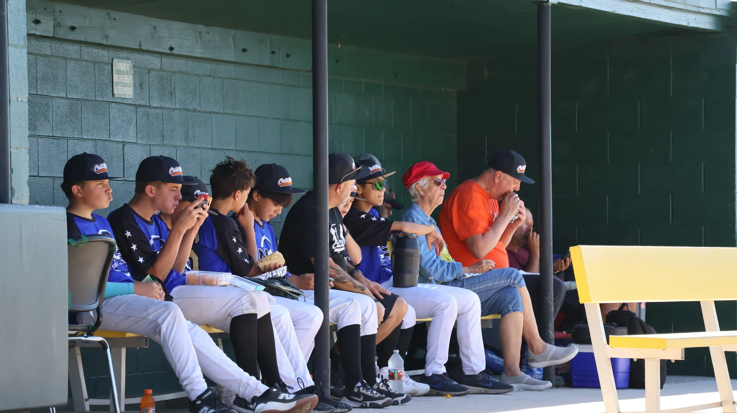 Baseball players in blue and black uniforms sitting in a dugout, with older spectators; some players are using phones, some are eating, and one appears to be drinking from a water bottle. The dugout has a dark green wall and a yellow bench.