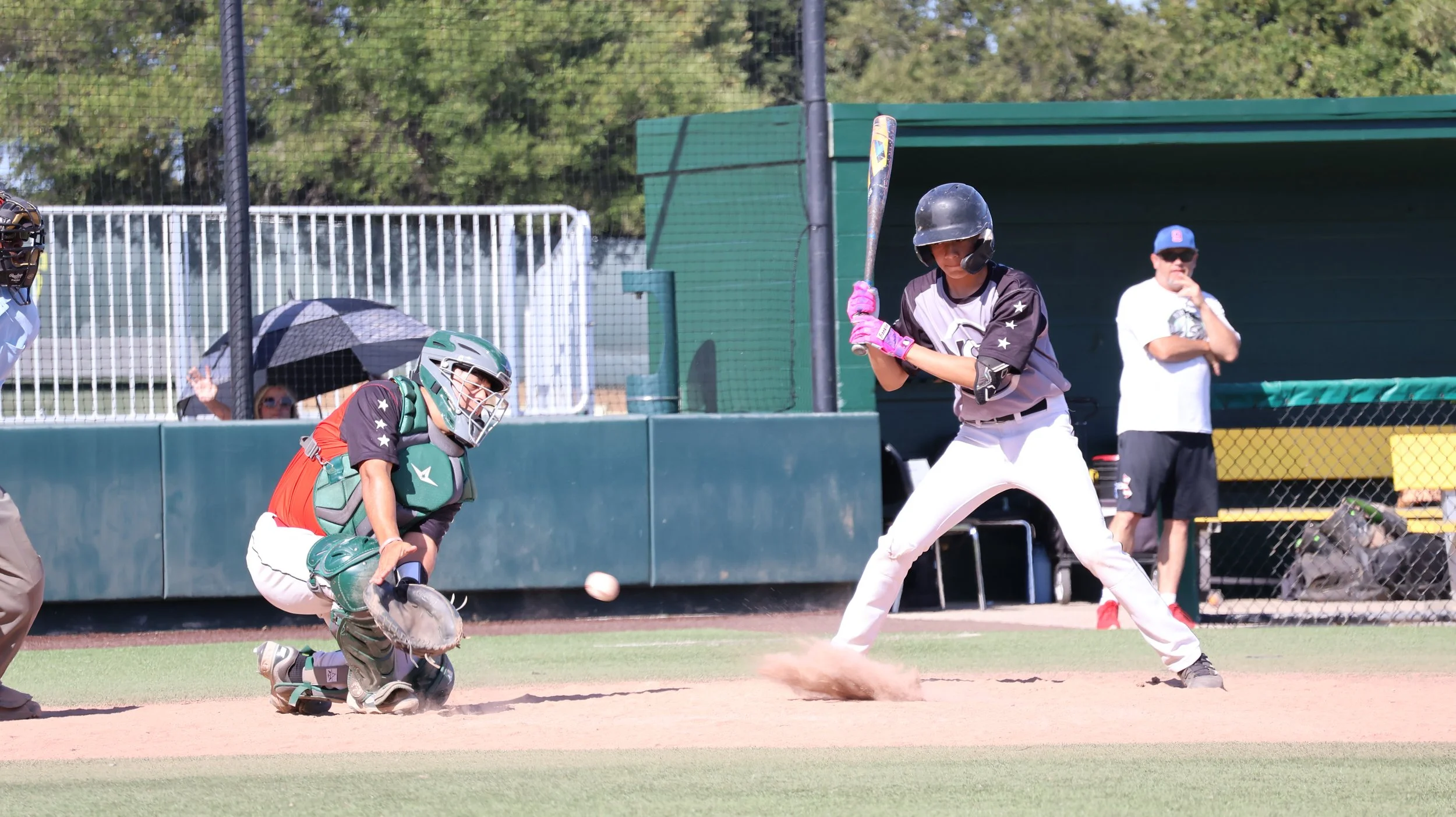 A baseball game scene with a batter in batting stance, a catcher crouched behind home plate, and a coach or spectator standing with arms crossed nearby. The batter wears a black helmet and gray uniform, holding a bat, while the catcher wears protecti