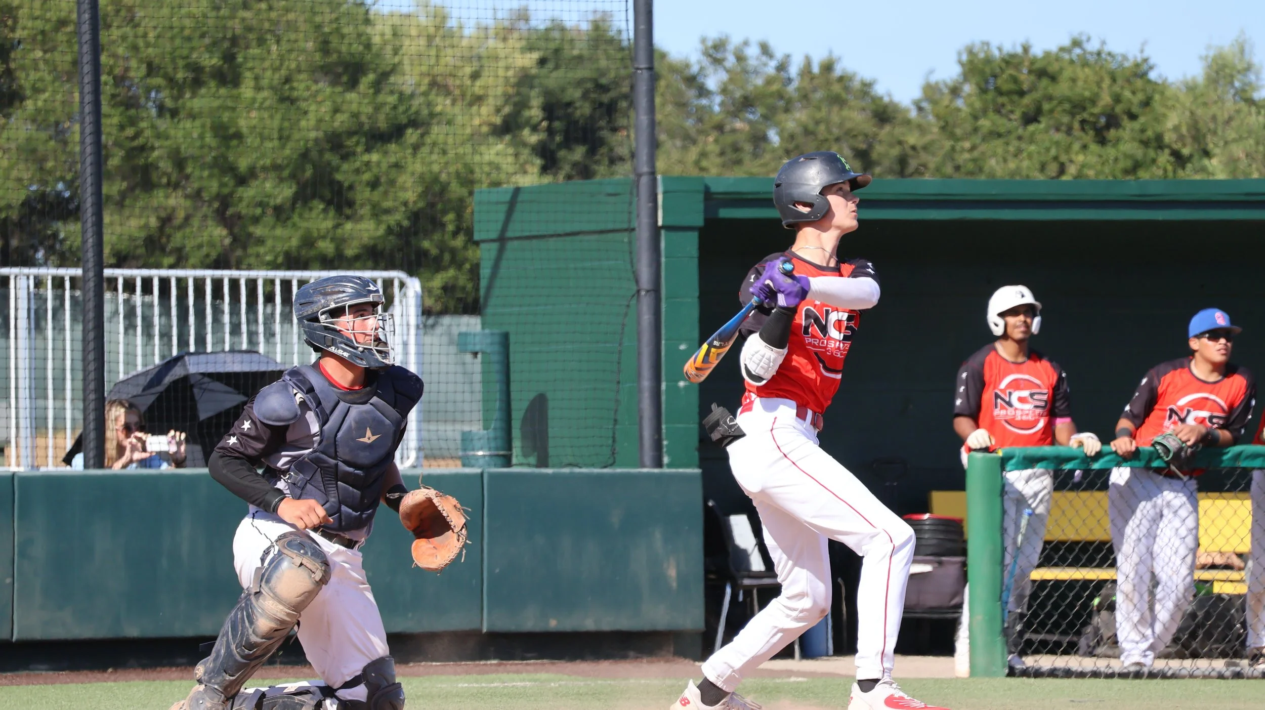 A baseball player swings at a pitch while the catcher behind him prepares to catch, with other team members and spectators watching from the dugout and behind the fence.