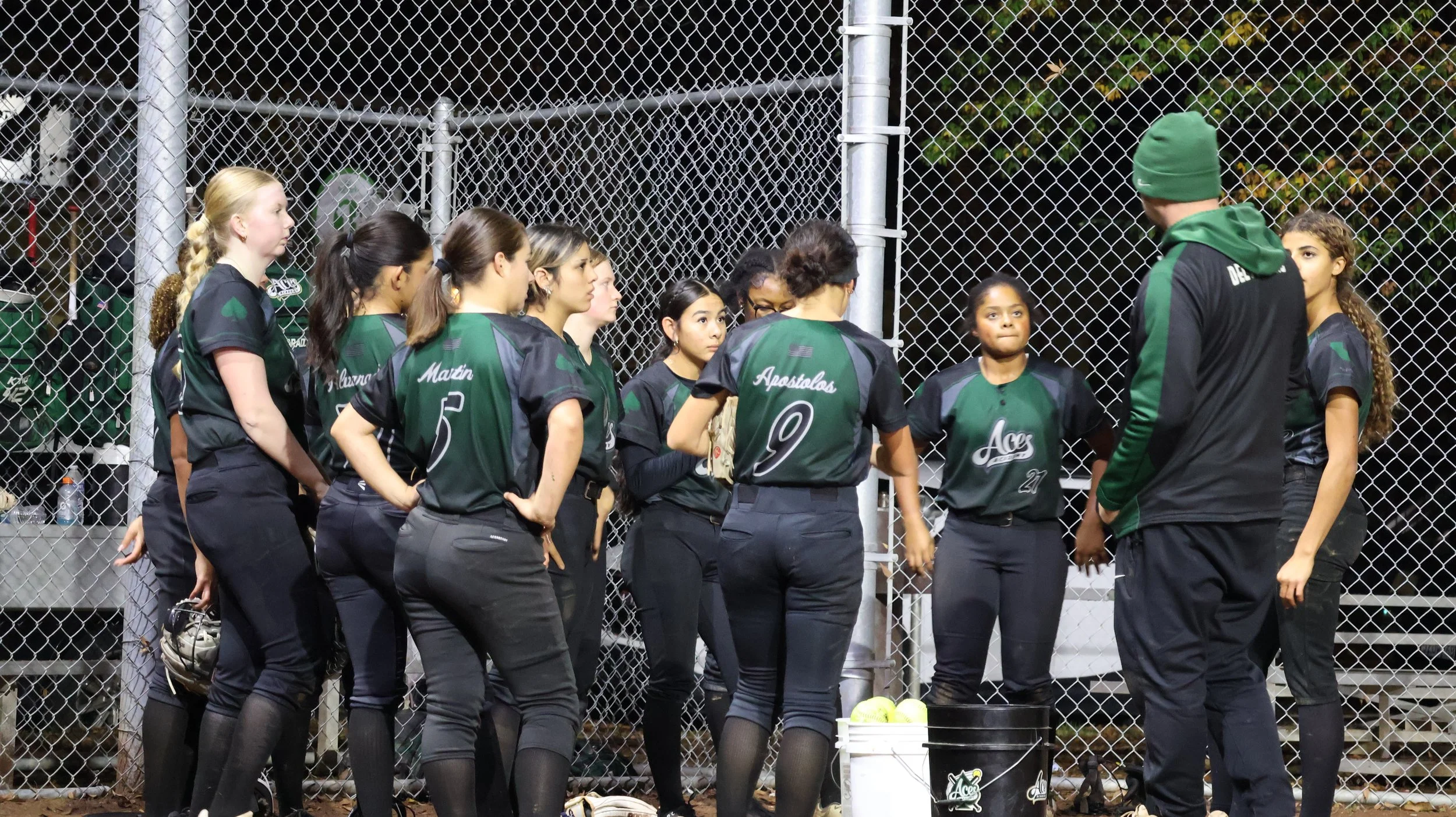 Girls softball team standing in a huddle around their coach on a field at night, wearing black and green uniforms.