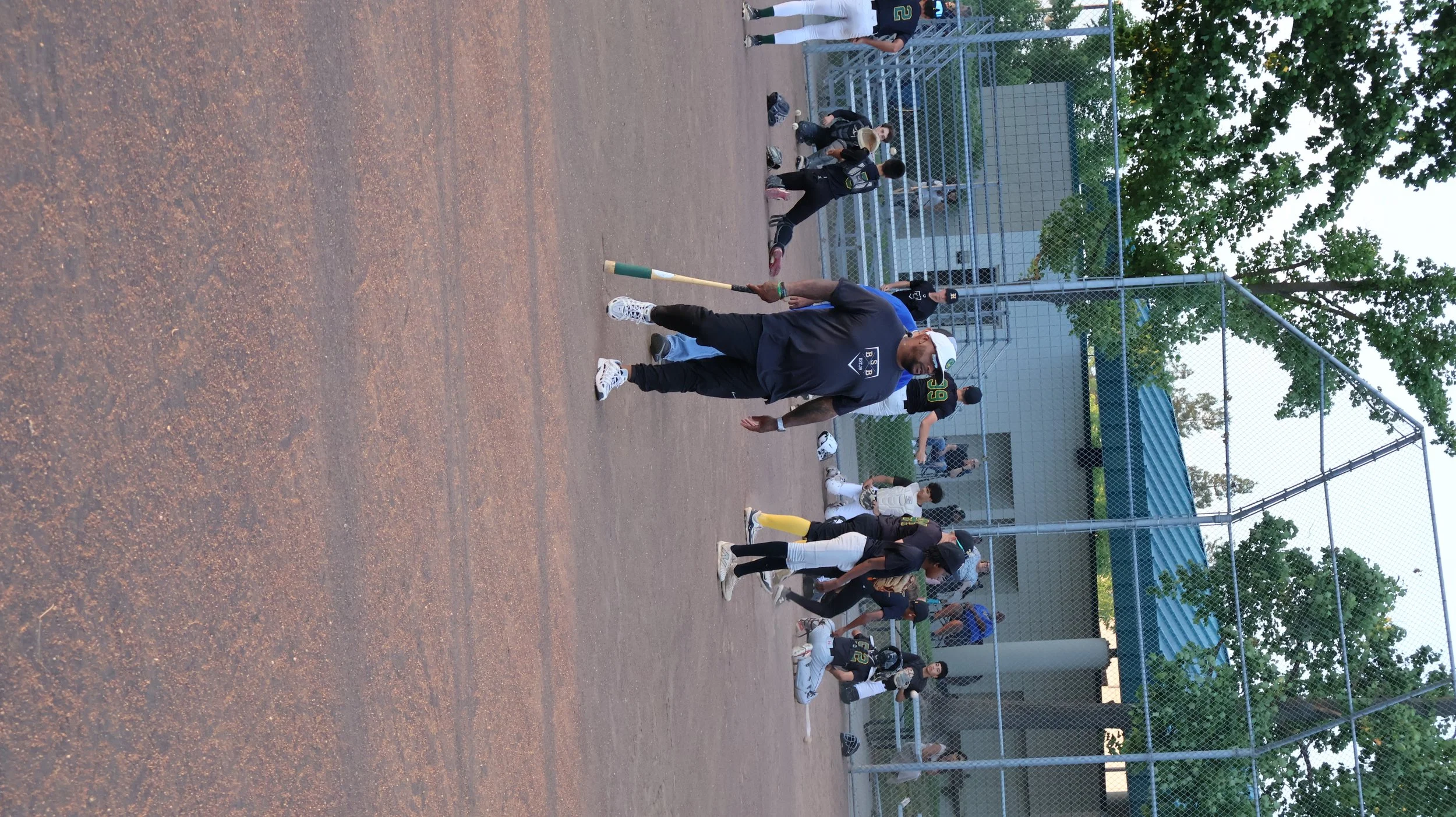 People playing baseball on a dirt field surrounded by a chain-link fence, with some sitting on the ground and others near the fence, under trees and a building in the background.