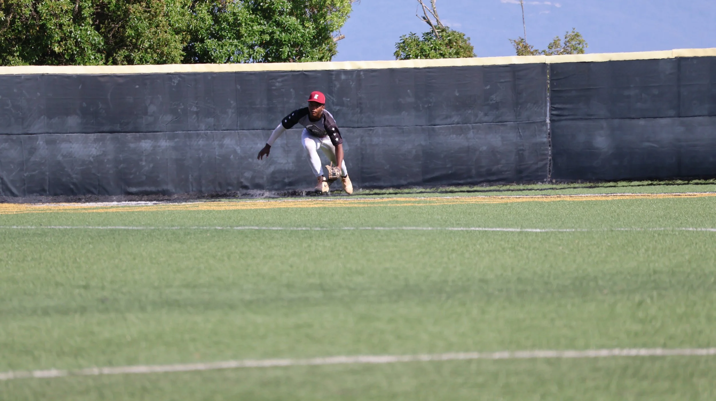 A baseball player in a black and white jersey, white pants, and a red cap is attempting to catch a ball near the outfield fence on a grassy field.
