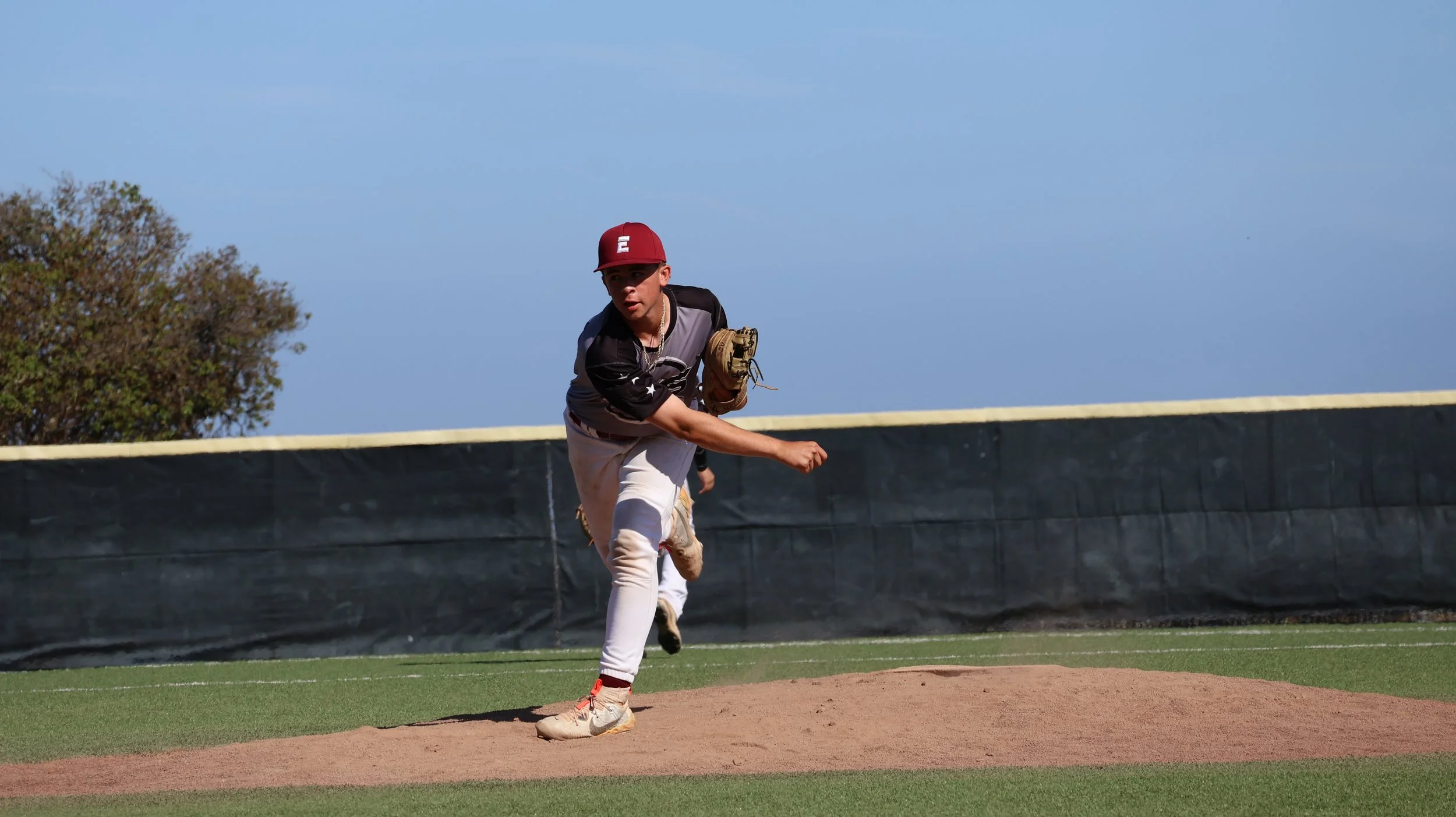 A baseball player in a black and gray uniform is pitching on the mound, wearing a red cap, white pants, and tan cleats. He is in a follow-through motion after throwing the baseball on a baseball field with a grass outfield and a blue sky.
