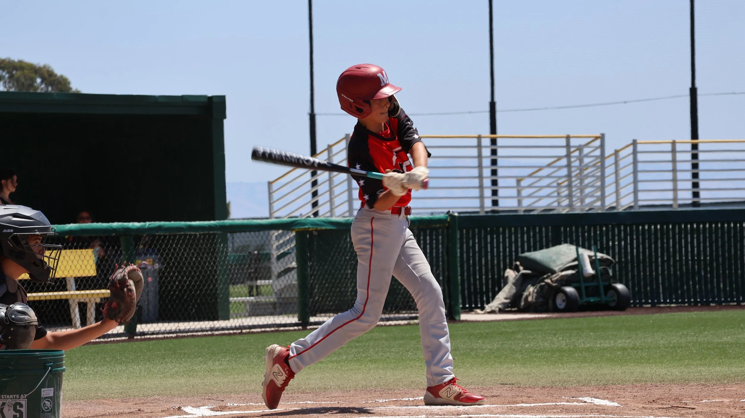 A young baseball player in a red helmet and black and red jersey is batting at a baseball game on a sunny day. The batter is swinging a black bat, with an umpire behind home plate in a black mask and uniform, and a catcher in black protective gear an