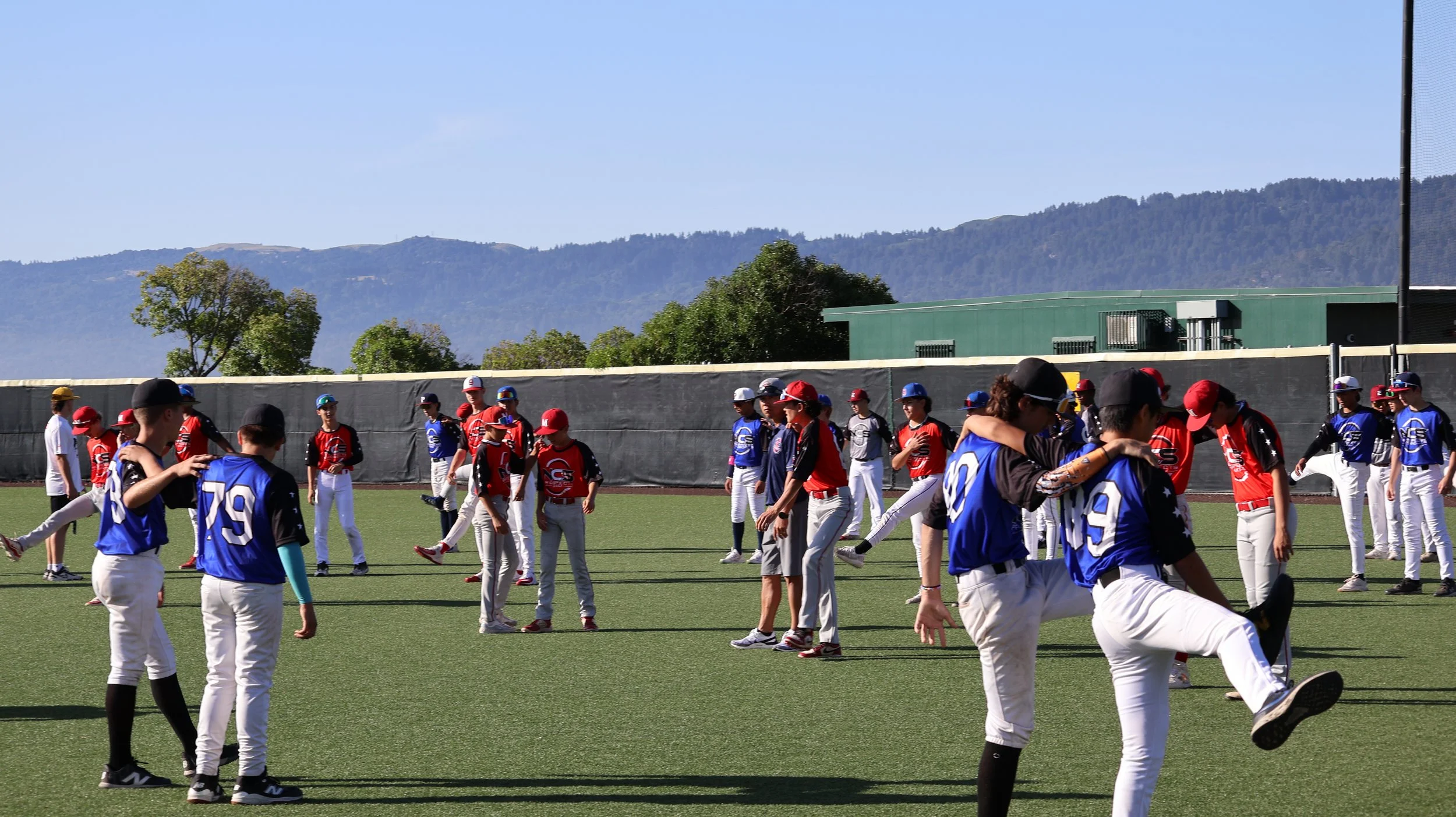 Youth baseball team practicing on field with mountains in background, players in red and blue uniforms engaging in drills and warm-up exercises.