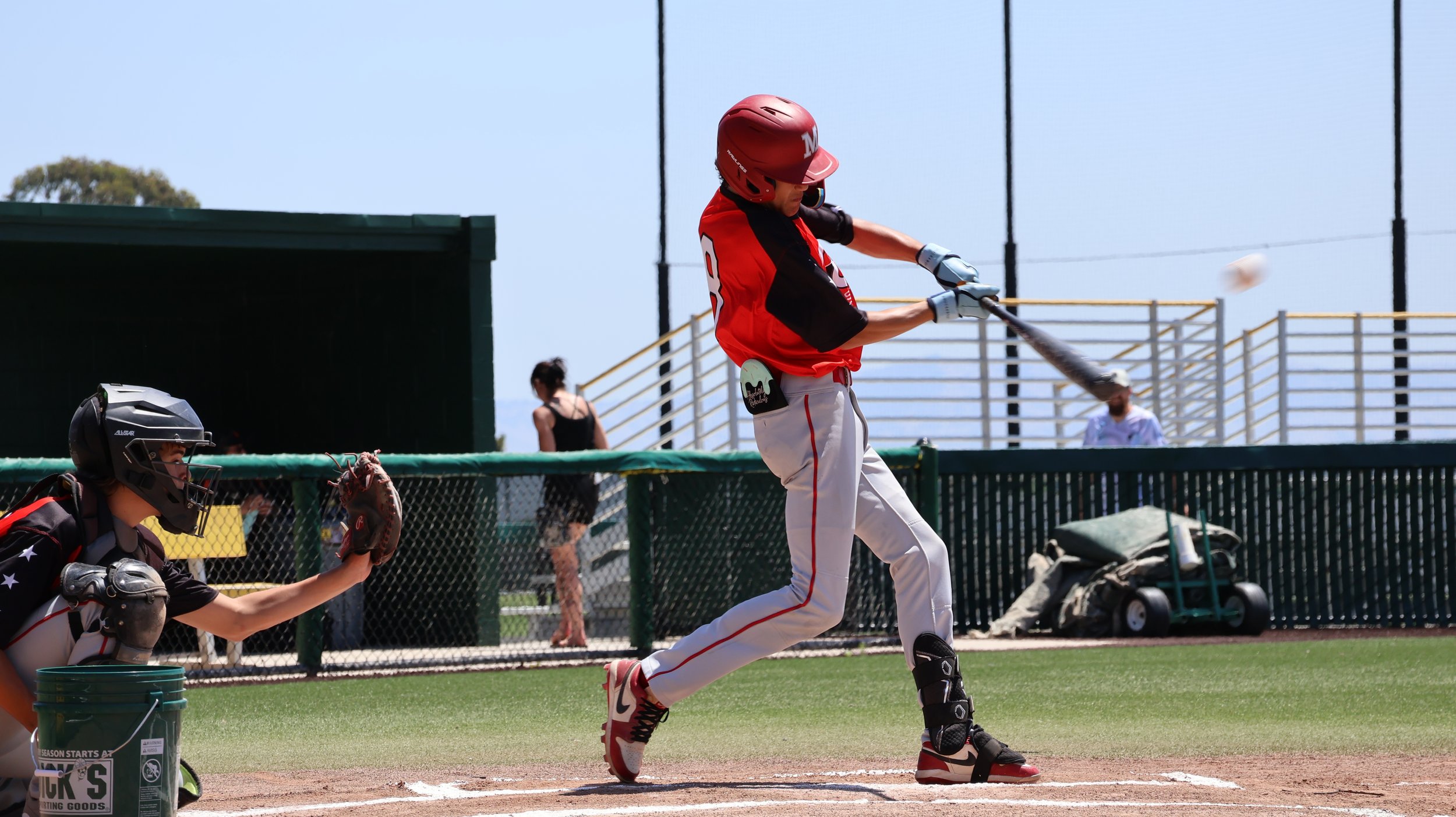 A baseball player in a red helmet and jersey swings a bat at a pitched ball on the field. The catcher in black protective gear crouches behind home plate, ready to catch. The scene takes place on a sunny day at the baseball field.
