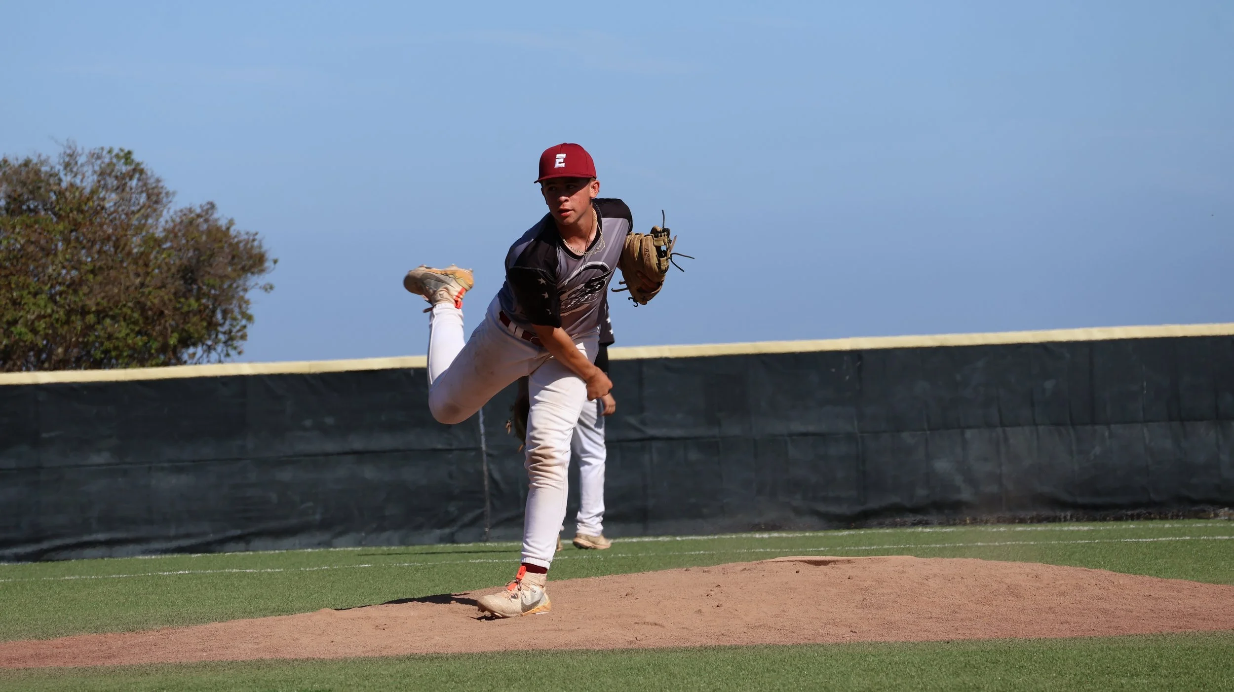 A young male baseball player in a red cap, black and gray jersey, and white pants, is pitching on a baseball field during daytime. He is in the middle of a pitch, with his right leg raised and his right arm extended forward.