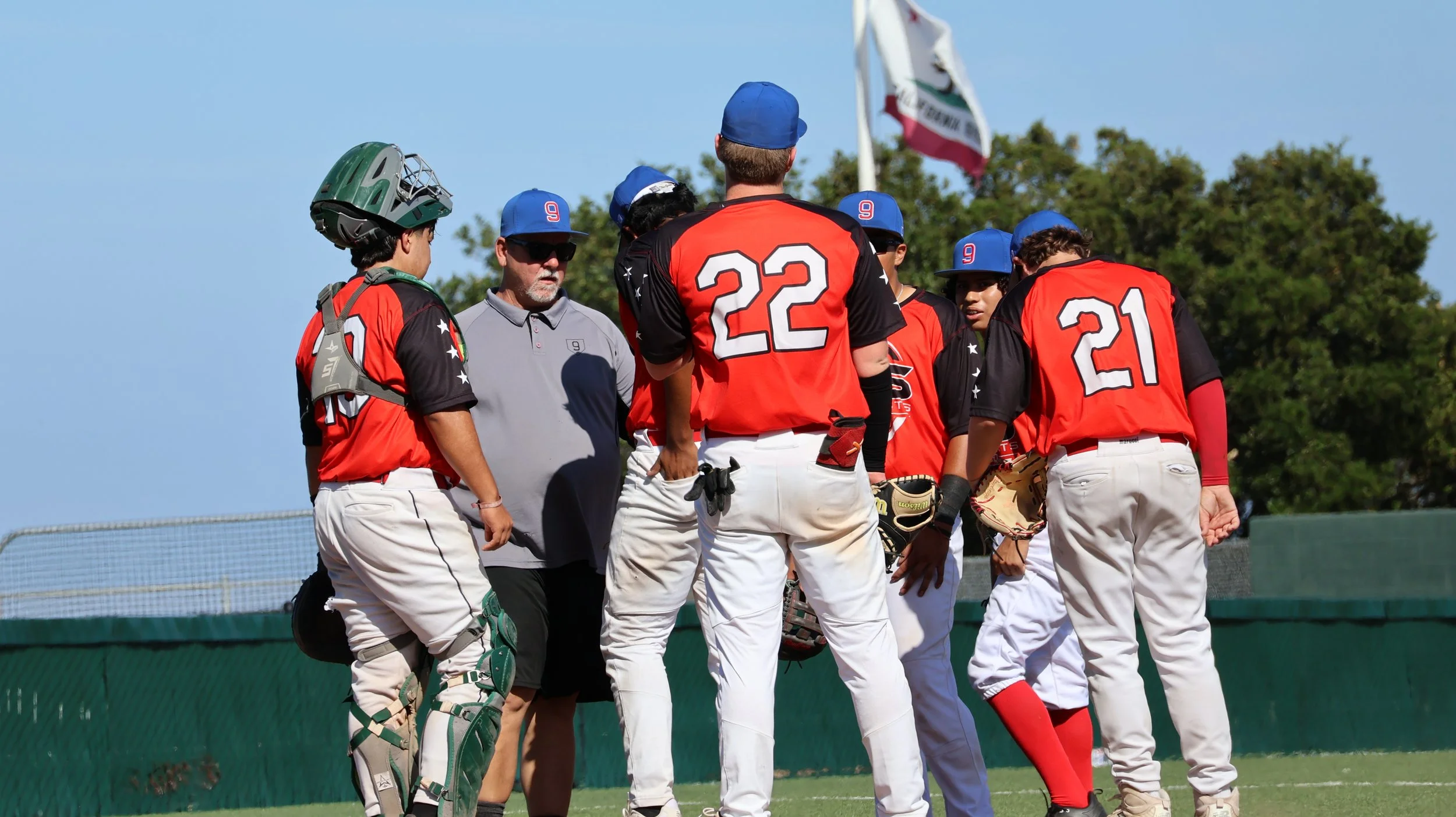Group of young baseball players and coach in huddle on the field with trees and blue sky in the background.