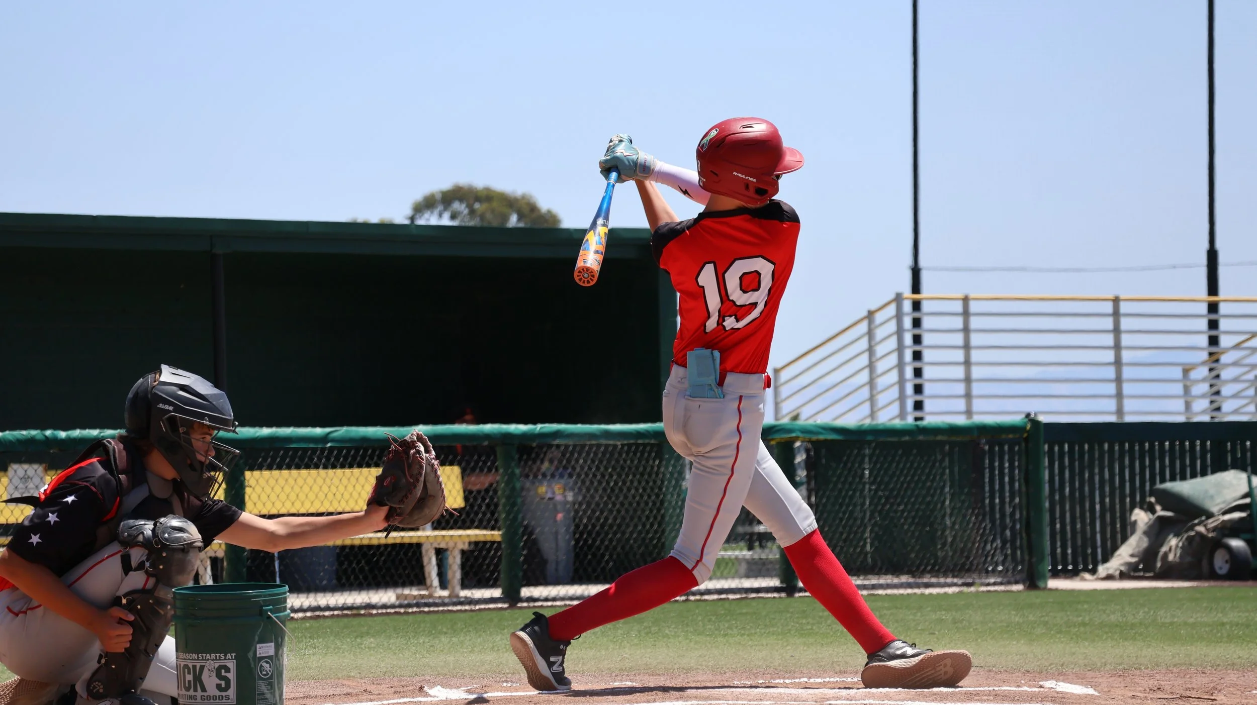 A young baseball player in a red jersey with number 19, red socks, and a helmet swings a bat at a pitched ball during a game on a baseball field.