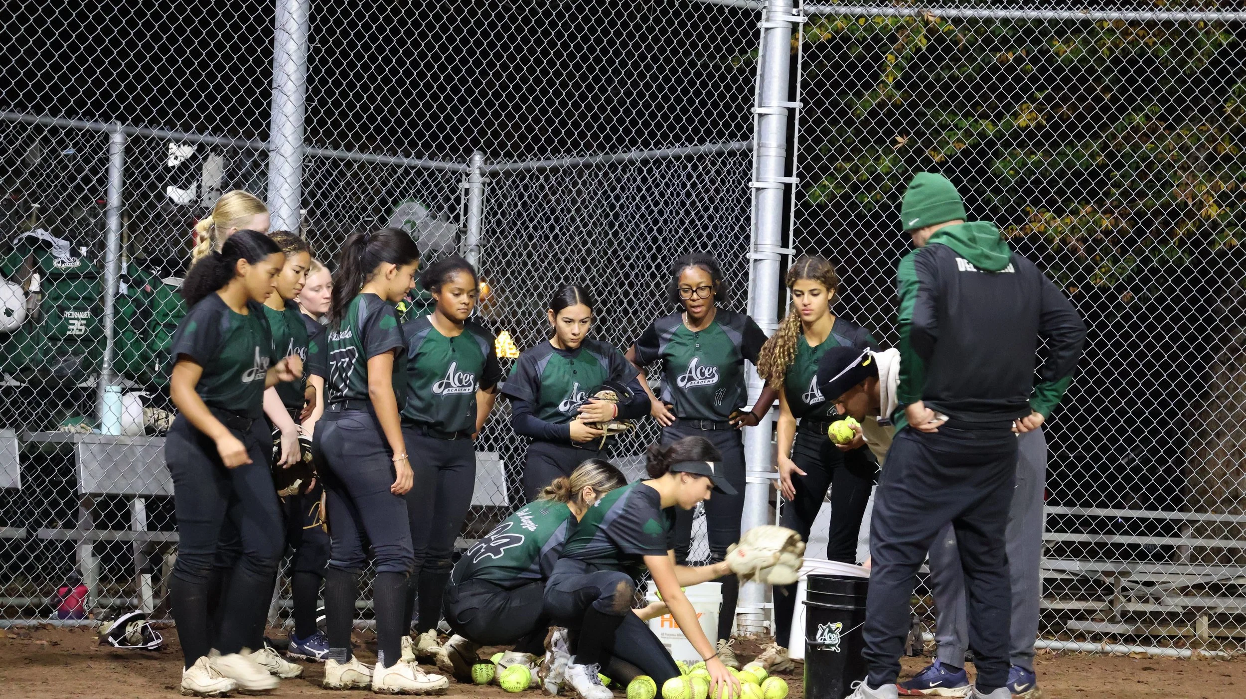 A women's softball team gathered on a field during a game or practice, with one player crouching down to pick up a ball, and a coach giving instructions. The team is wearing matching green and black jerseys and black pants, standing near a chain-link