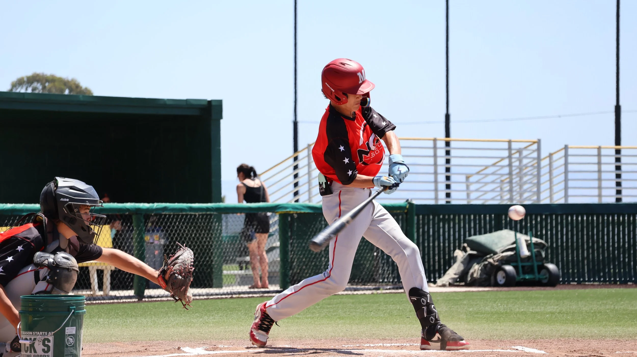 A baseball player in a red helmet and jersey hits a pitched ball during a game, while the catcher in black gear crouches nearby, on a baseball field with green grass and a chain-link fence.