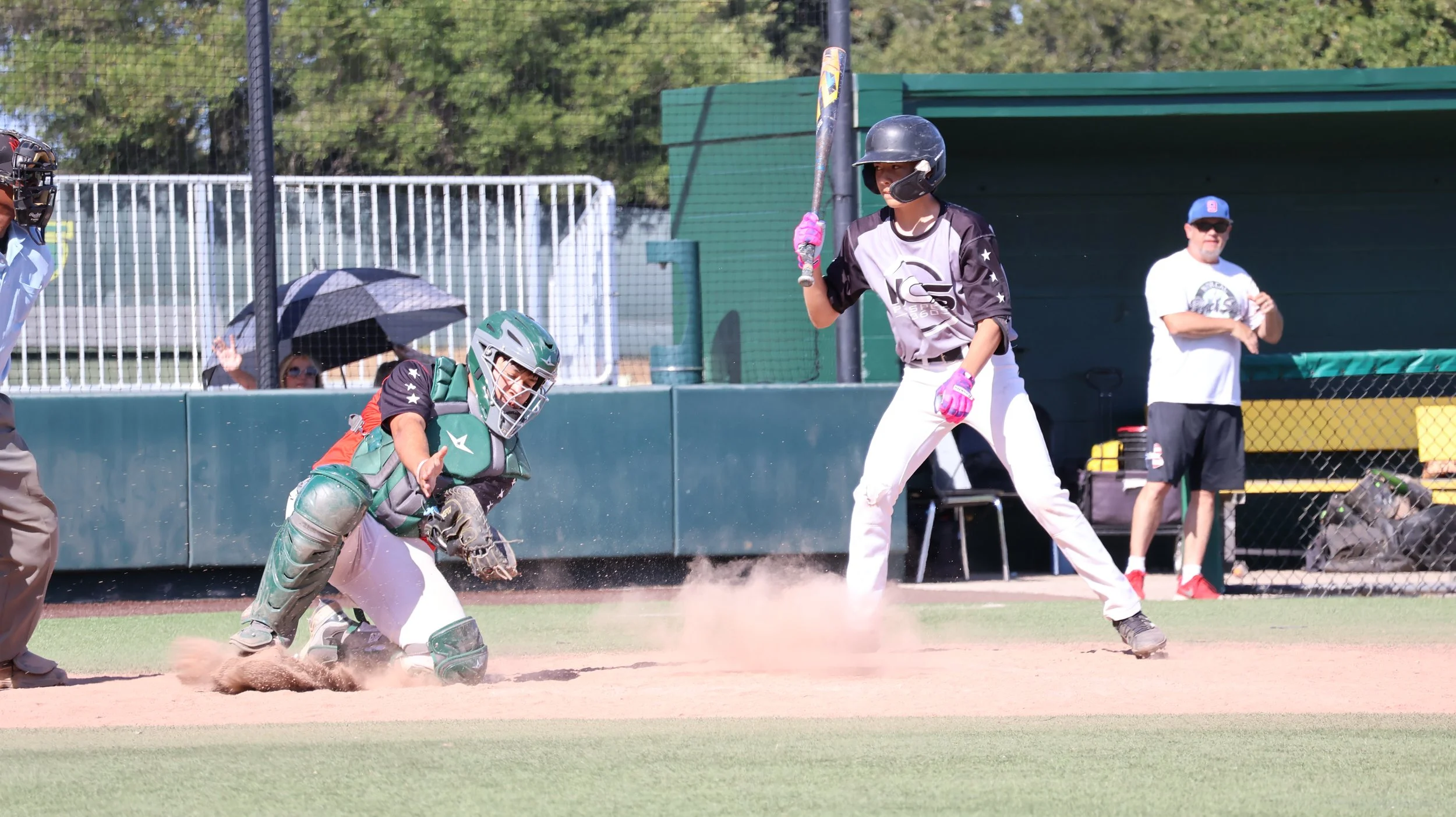 Baseball player at home plate preparing to swing, catcher crouching behind him, umpire to the side, and coach or team staff standing in the background near the dugout, with a person holding an umbrella in the stands.