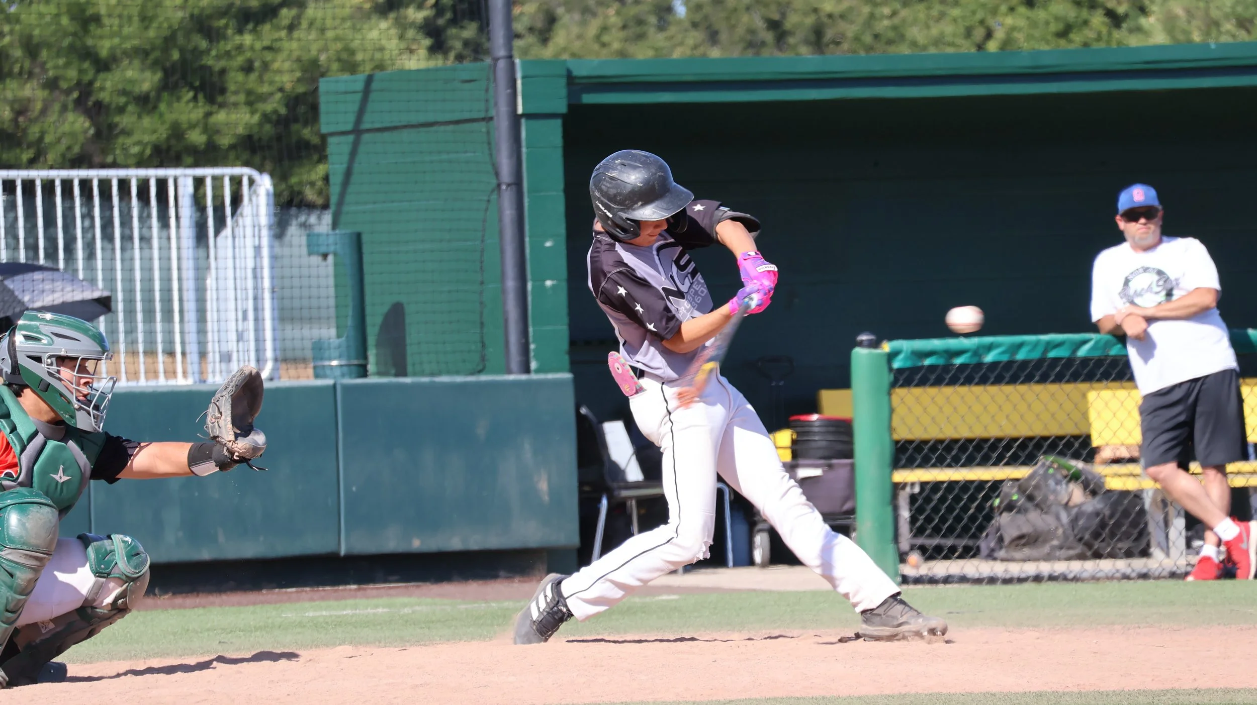 A baseball player swinging a bat at a pitched ball on the field. The catcher is crouched behind home plate, and an observer stands in the dugout area observing the game.