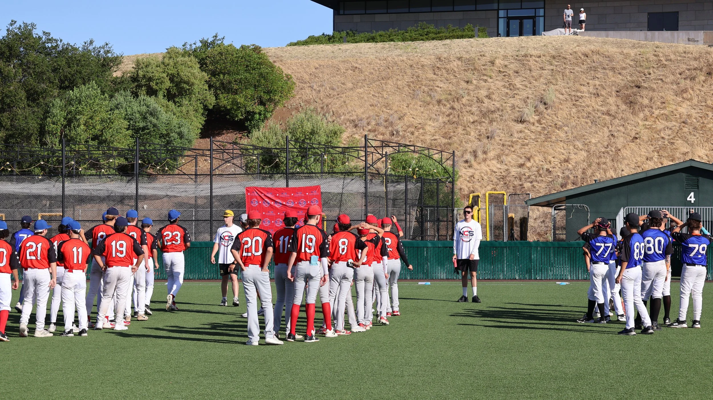Youth baseball teams in red and blue uniforms standing on the field facing each other, with coaches or officials nearby, some kids adjusting their caps, on a sunny day.