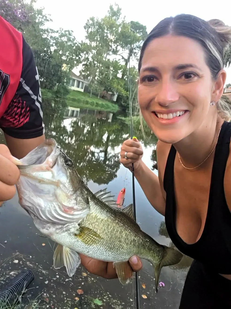 Woman smiling while holding a large fish she caught during a fishing trip by a lake.