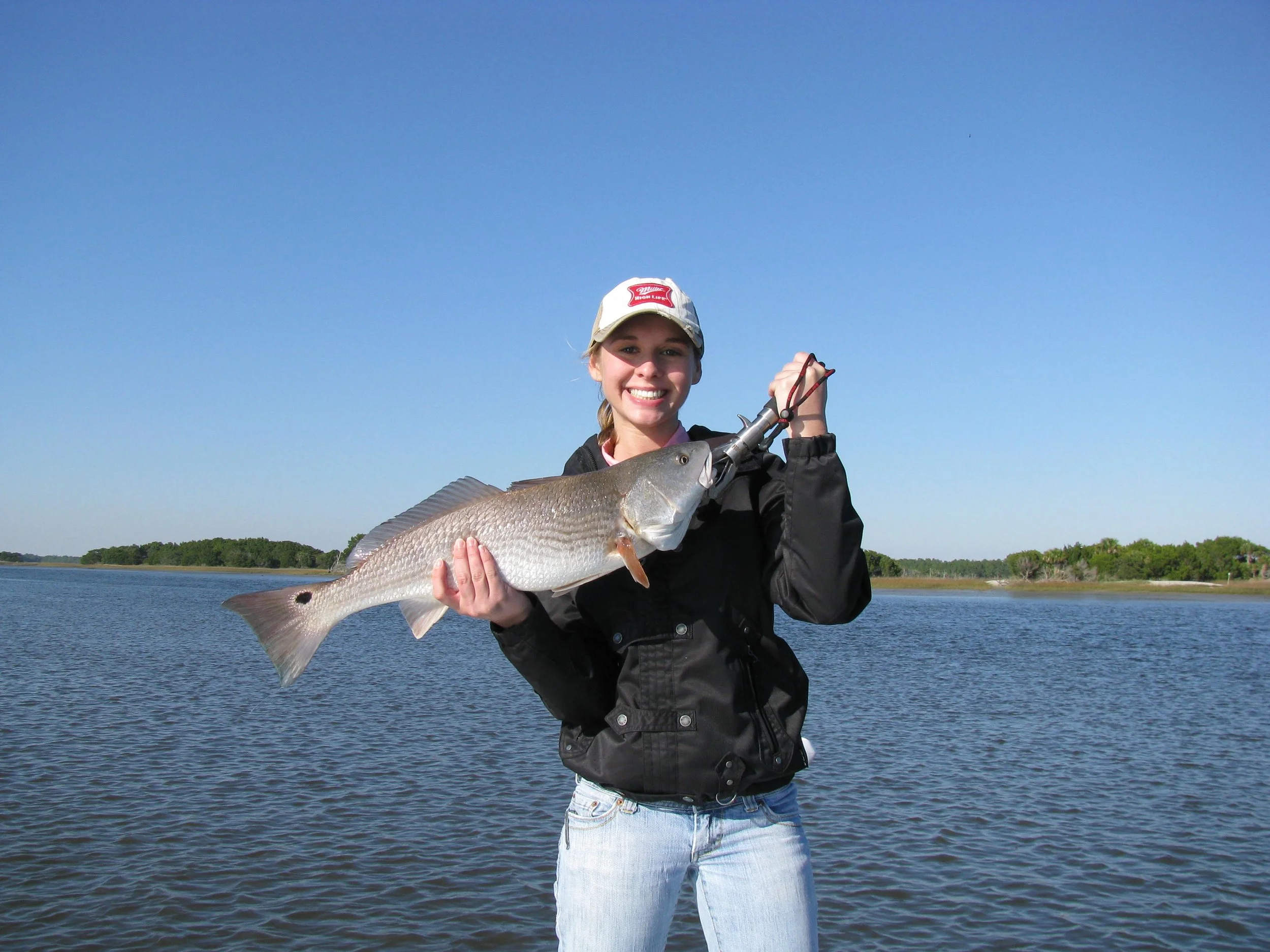 A young woman in a black jacket and a white cap holding a large fish on a fishing line, standing by a river on a sunny day with a clear blue sky.