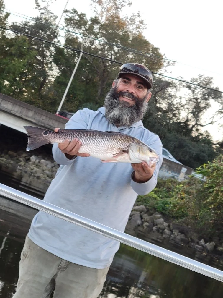 A man with a beard, wearing a long-sleeve shirt and a cap, holding a large fish outdoors near a river or lake, smiling at the camera.