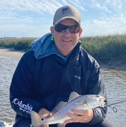Man in outdoor fishing gear holding a fish on a riverbank