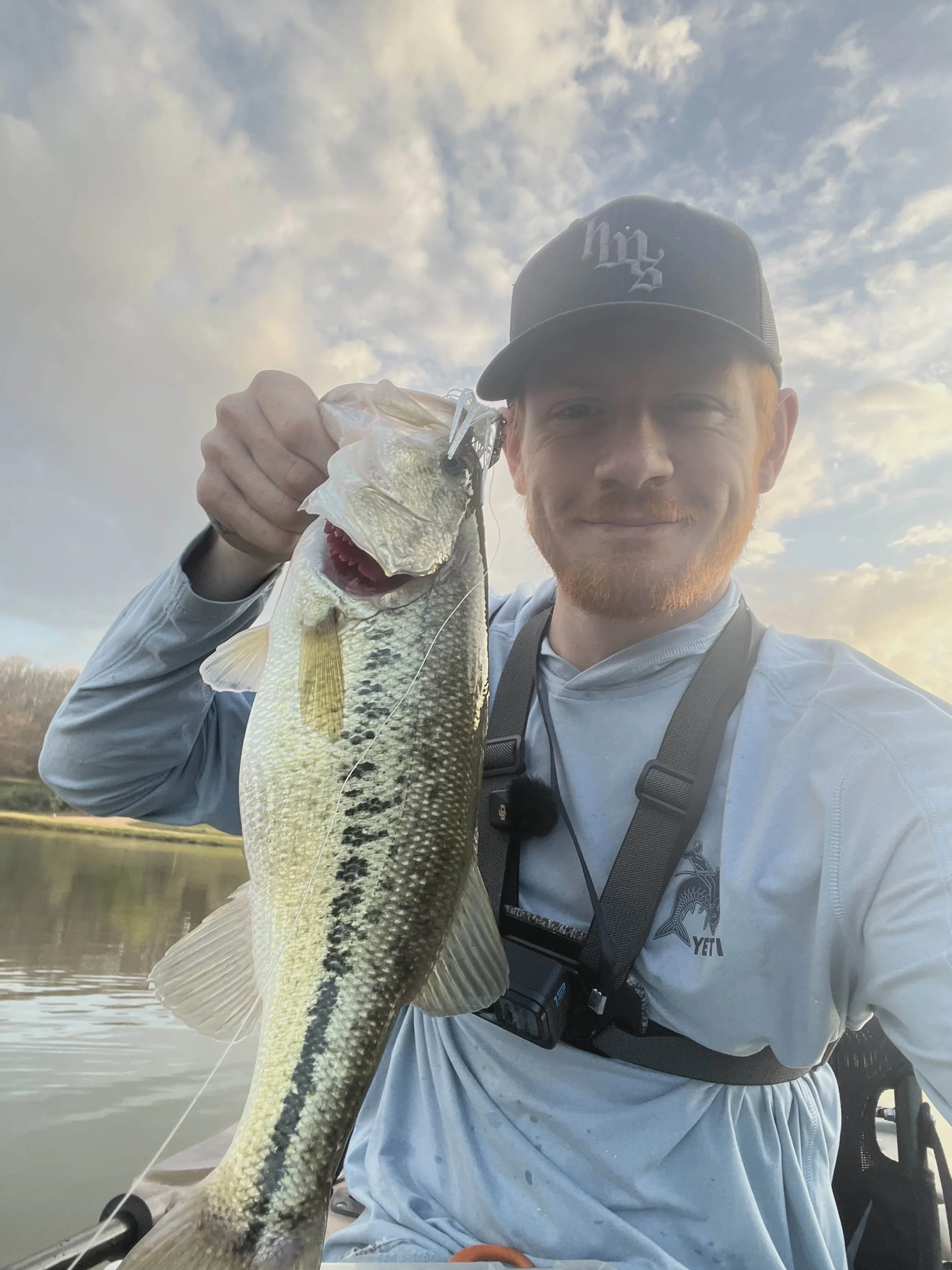 A man with a beard and wearing a black baseball cap is holding a largemouth bass fish. The man is wearing a white long-sleeved shirt and has a camera or device hanging around his neck. The background shows a body of water and a cloudy sky.