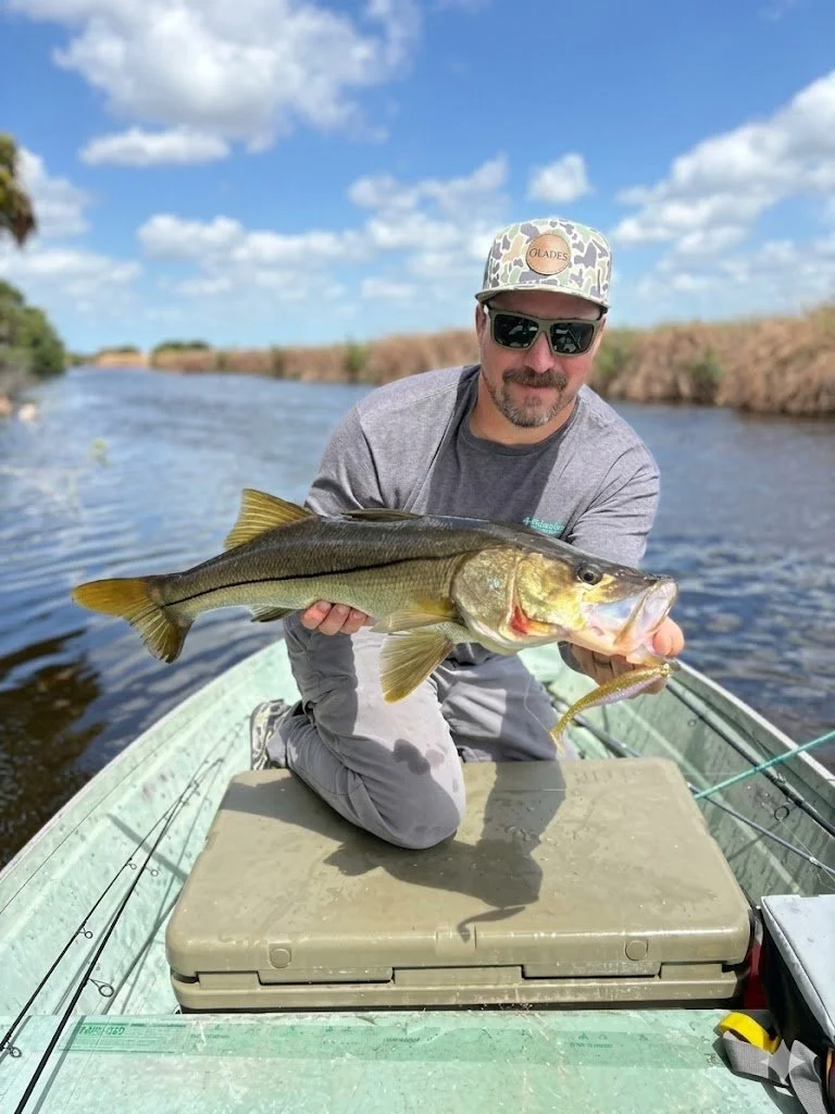 A man sitting in a small boat holding a large fish he caught, with water and blue sky in the background.
