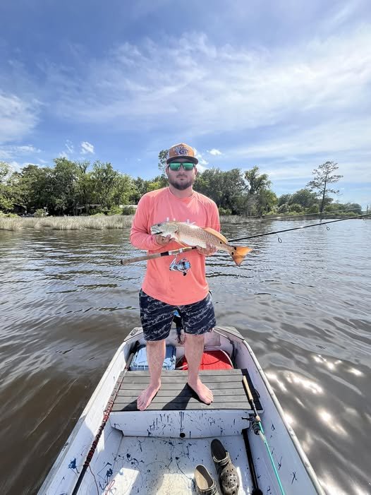 Man standing in a small boat on a river, holding a fish he caught, with blue sky and trees in the background.
