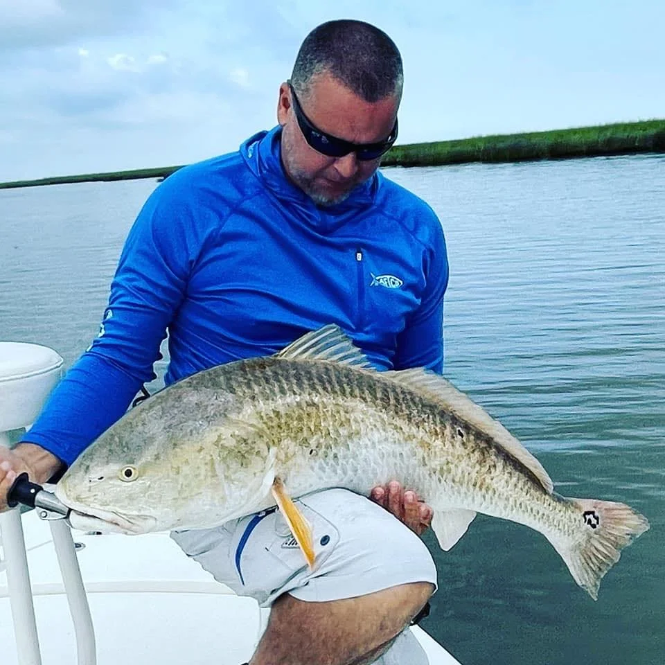 Charter Capt Armando in a blue shirt sitting on a boat, holding a large fish by the water, looking down at his catch.