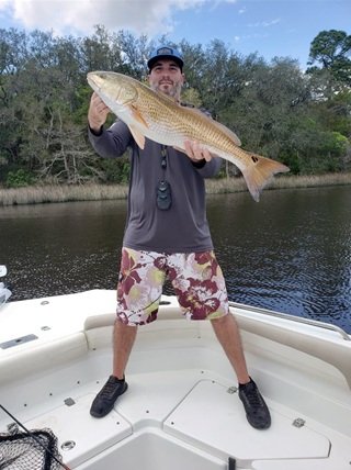 Man holding a large fish on a boat near a river, with trees in the background.