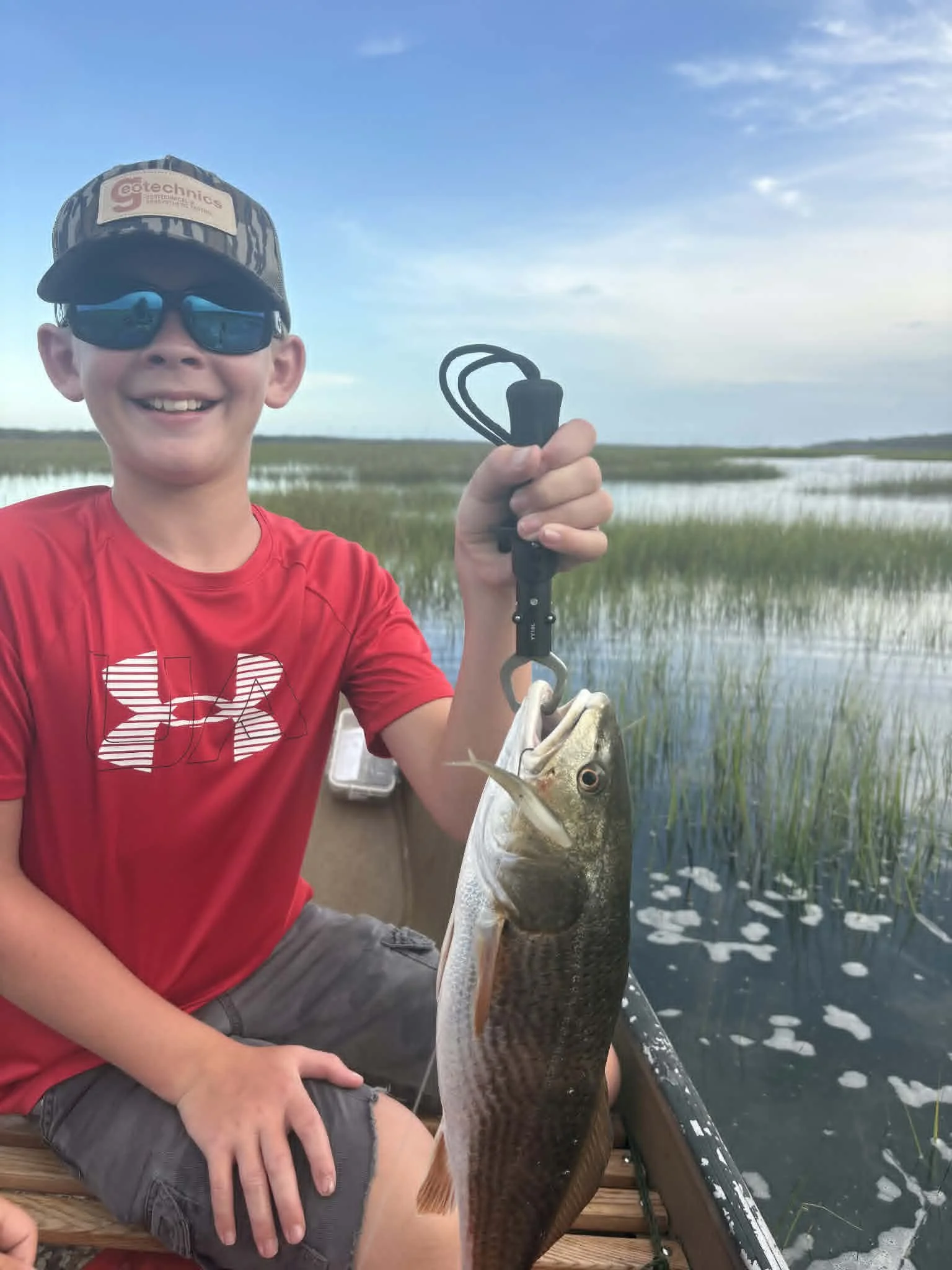 A boy smiling while sitting on a dock, holding a fishing rod with a fish caught on the line, in a marshy area with water and tall grass under a partly cloudy sky.