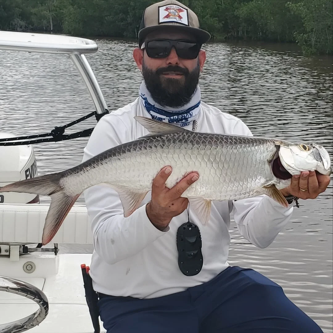 Man on a boat holding a large fish, wearing sunglasses, cap, white long sleeve shirt, and a neck gaiter, with water and trees in the background.