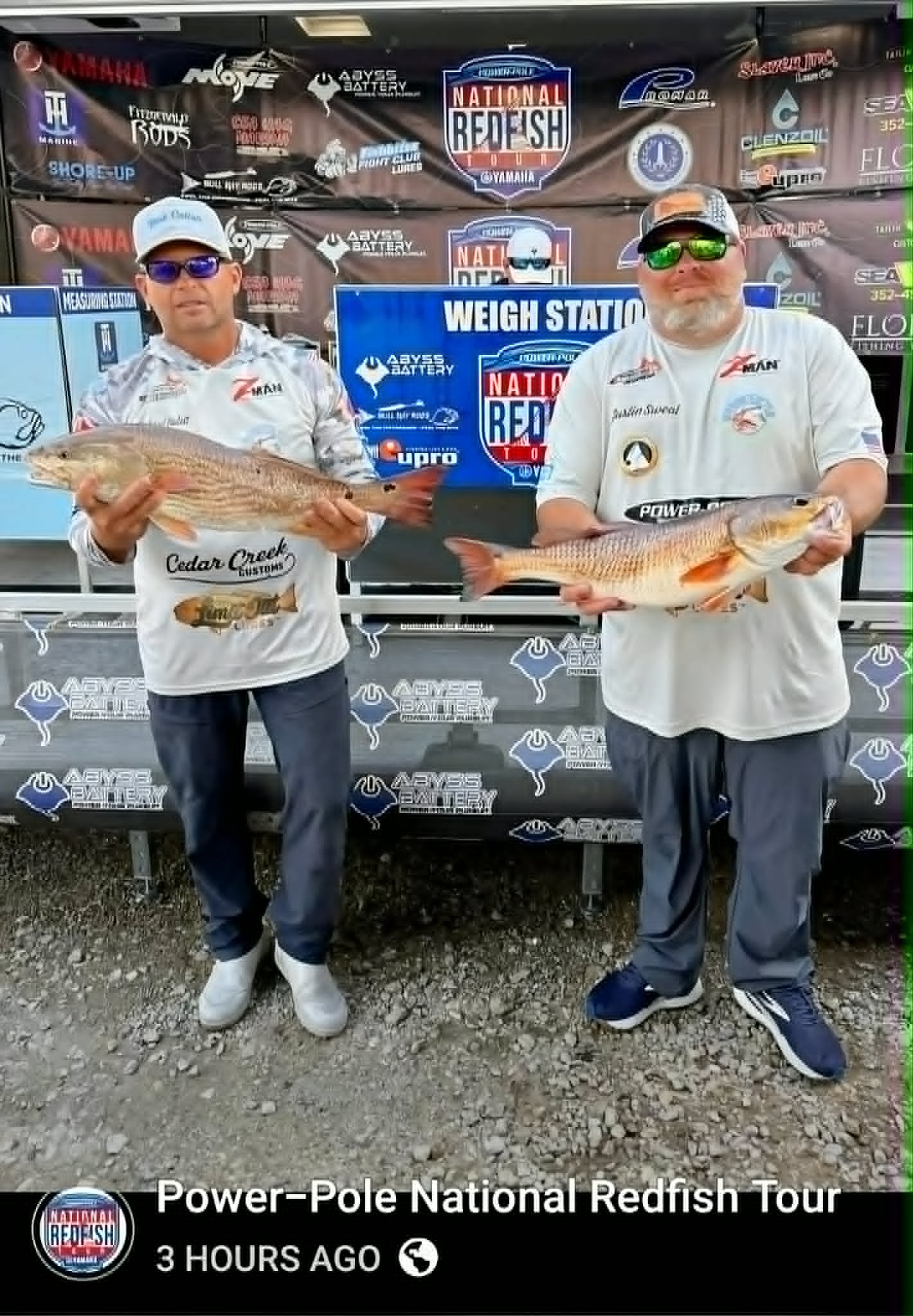 Two men in fishing gear holding large redfish in front of a branded backdrop at the Power-Pole National Redfish Tour, with a digital banner showing the event name, time since posted, and a globe icon.