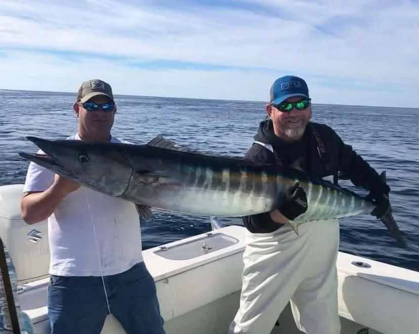 Two men on a boat holding a large fish they caught, with ocean and sky in the background.