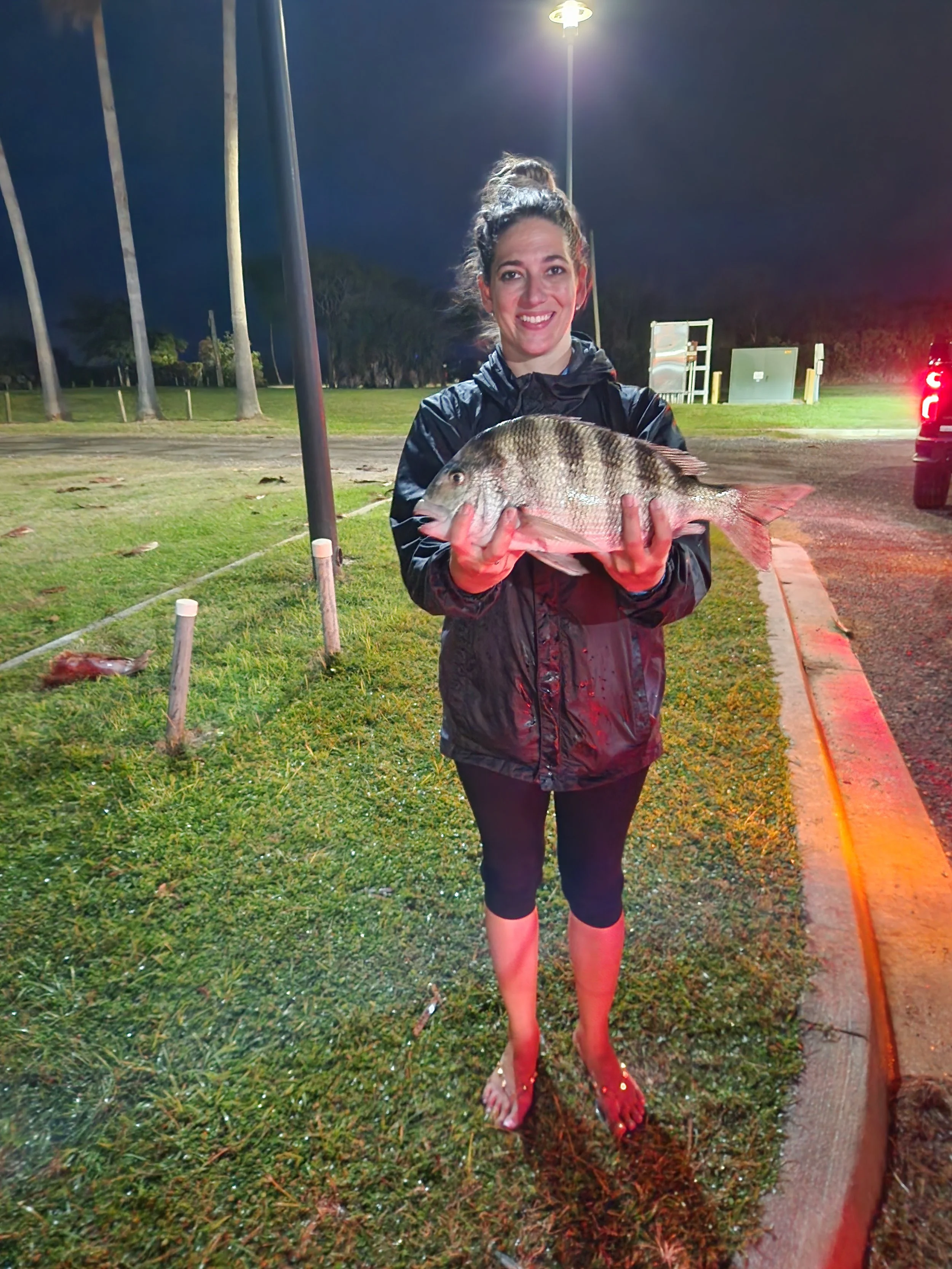 Woman standing outdoors at night, holding a large fish with multiple dark vertical stripes.