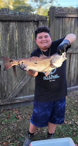 A young boy smiling and holding a large fish against a wooden fence in an outdoor setting.