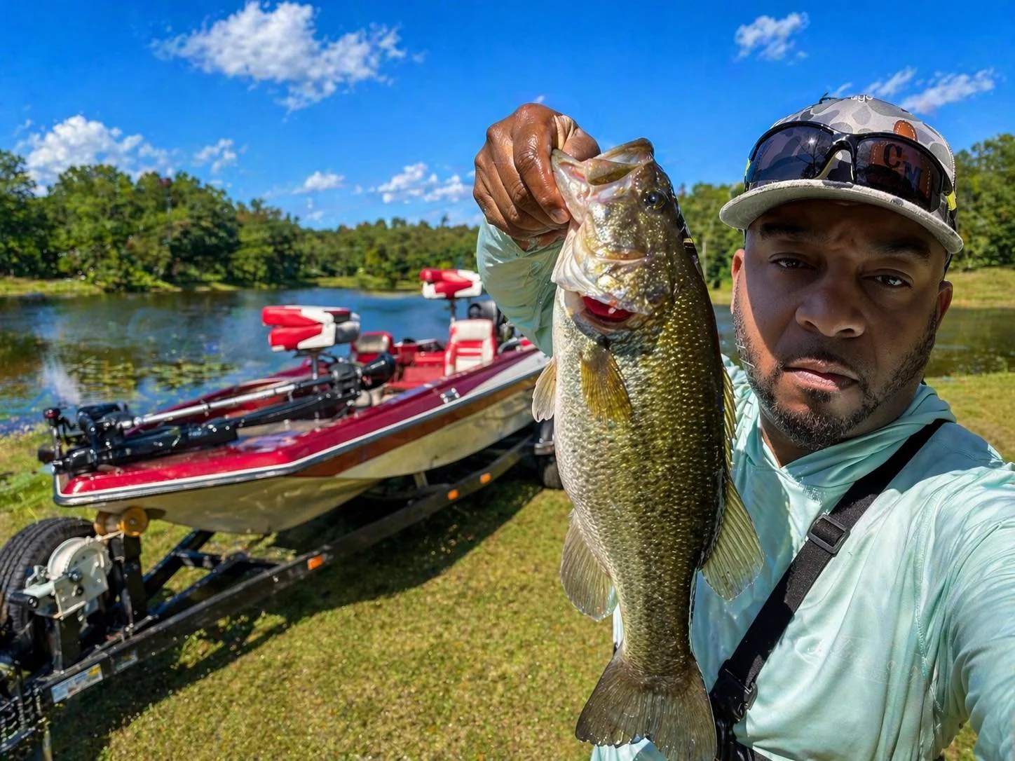 Man holding a large fish in front of a boat on a lake with trees in the background.
