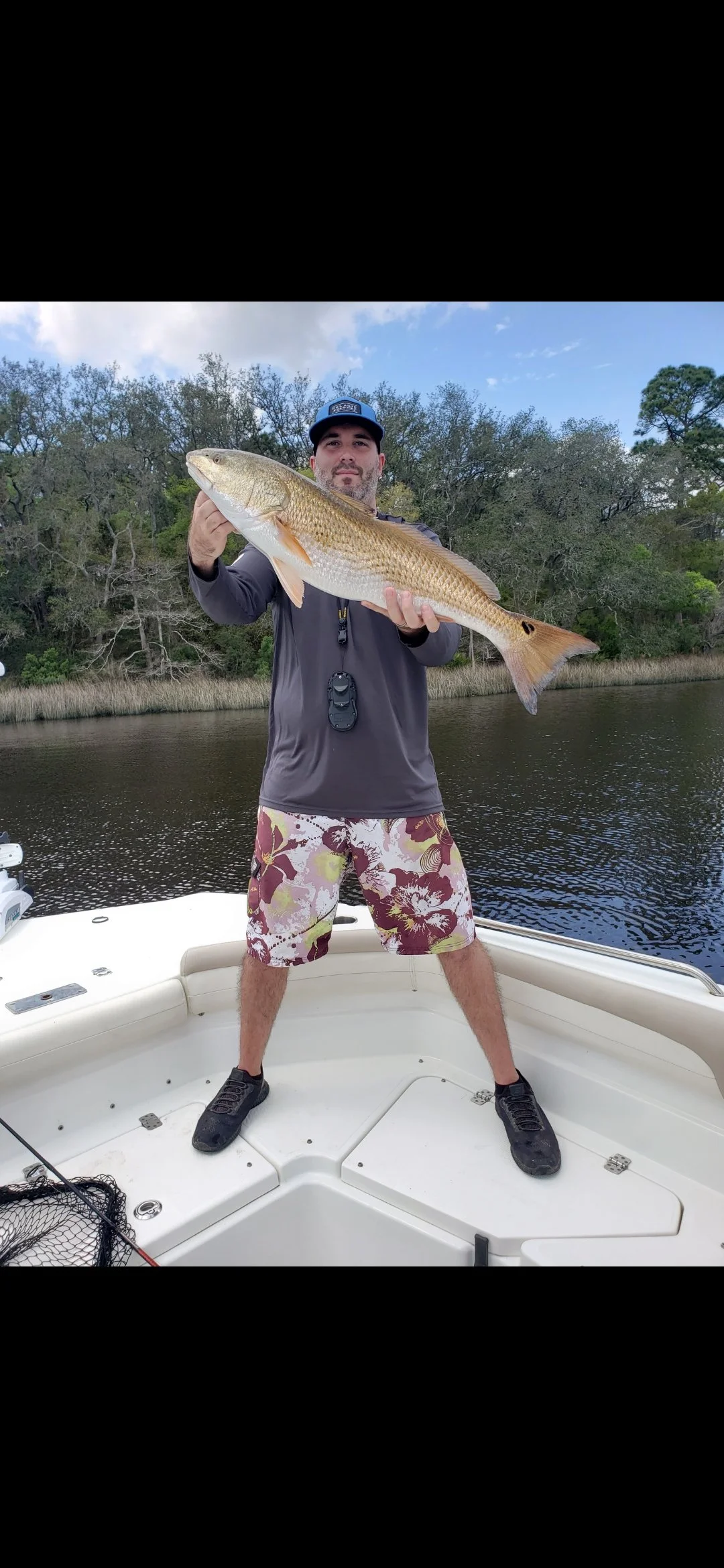 Man holding a large fish on a boat in a waterway with trees in the background.