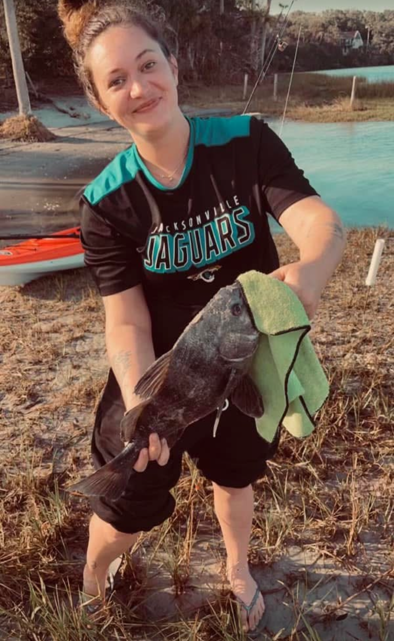 A woman with a high bun hairstyle wearing a Jacksonville Jaguars sports jersey is kneeling on a sandy, grassy area near water, holding a large fish with a green towel. A kayak and water are visible in the background.
