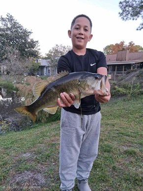 Young boy smiling and holding a large fish outdoors in a grassy area with trees and houses in the background.