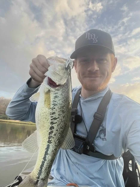Man in a cap and blue shirt holding a largemouth bass while fishing on a lake, smiling at the camera.