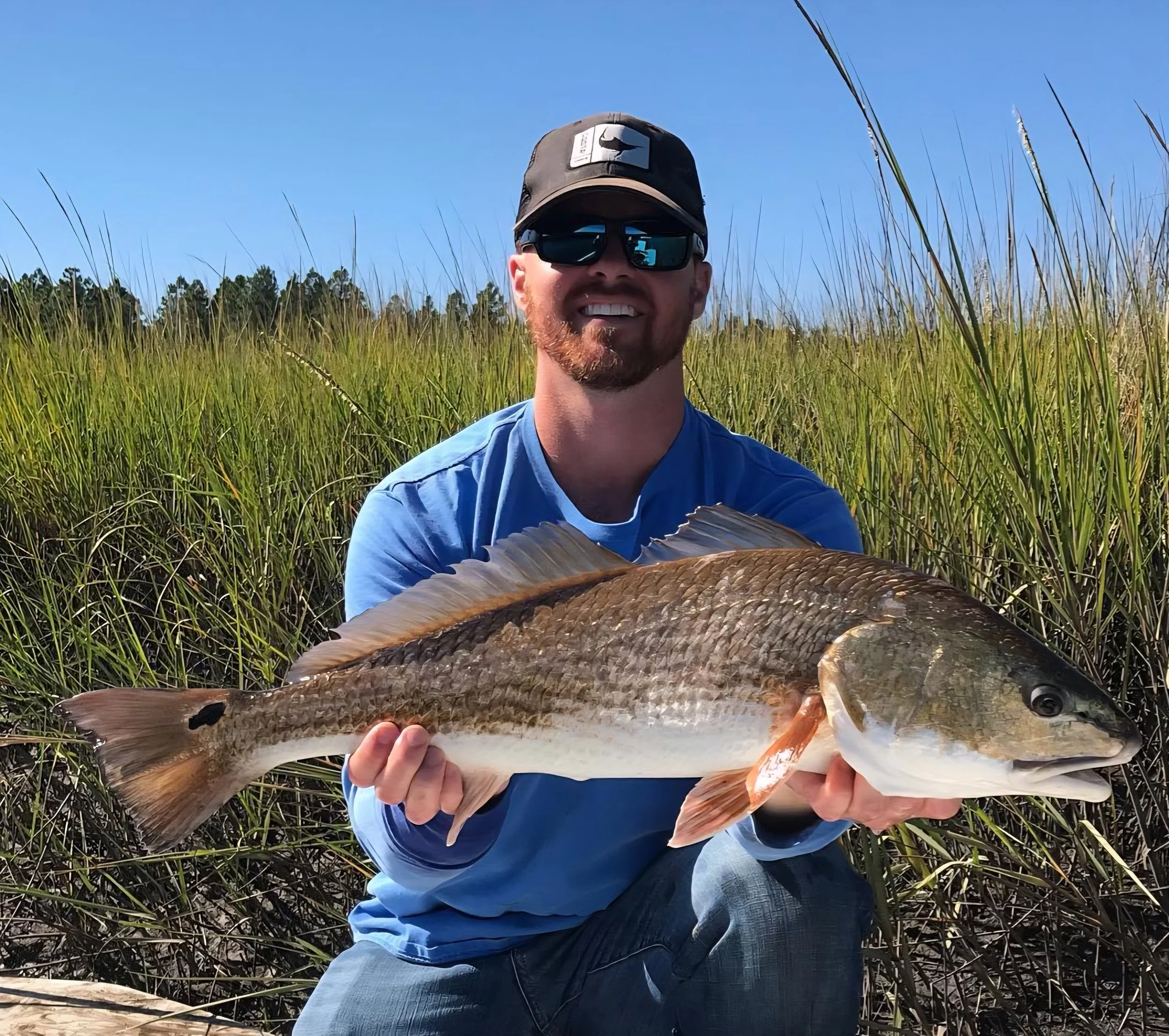 Man in sunglasses and a cap holding a large fish in a grassy marshland area with trees in the background.