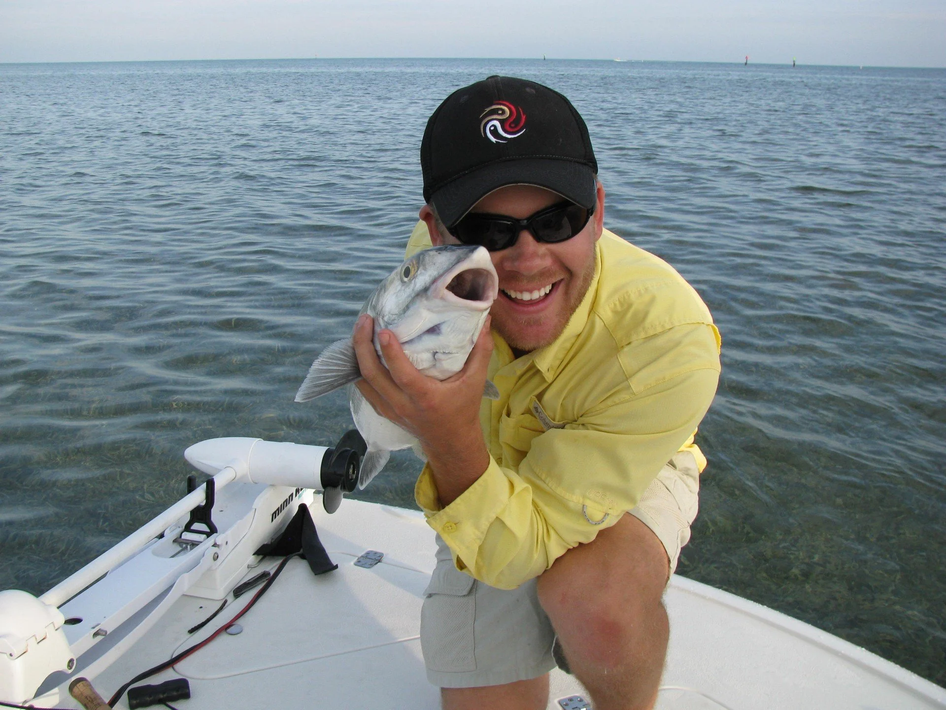 A man in a yellow shirt, sunglasses, and a black cap holding a fish on a boat in the water, smiling.