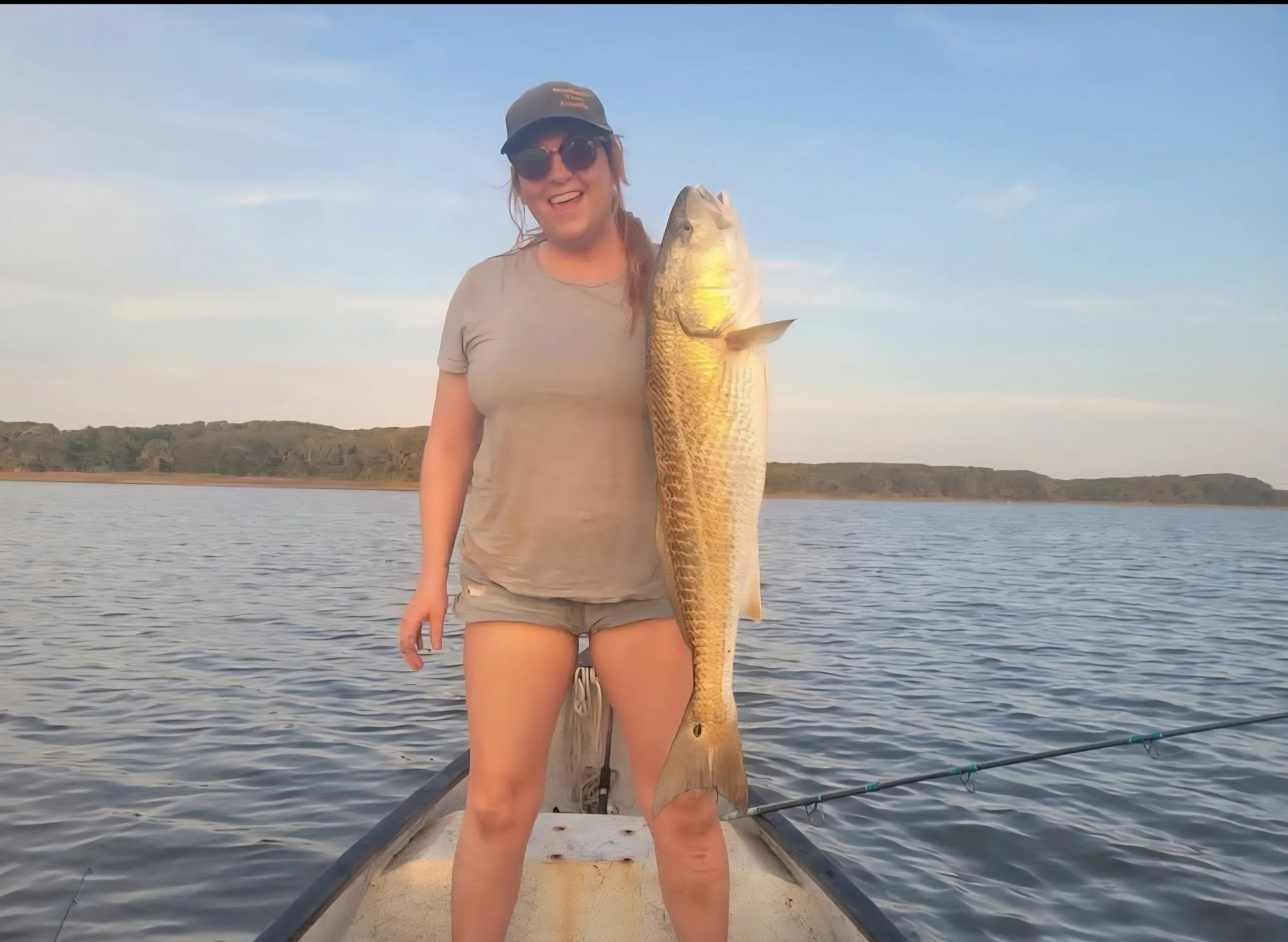 Woman smiling and holding a large fish on a boat in a body of water, with a distant shoreline and clear sky in the background.