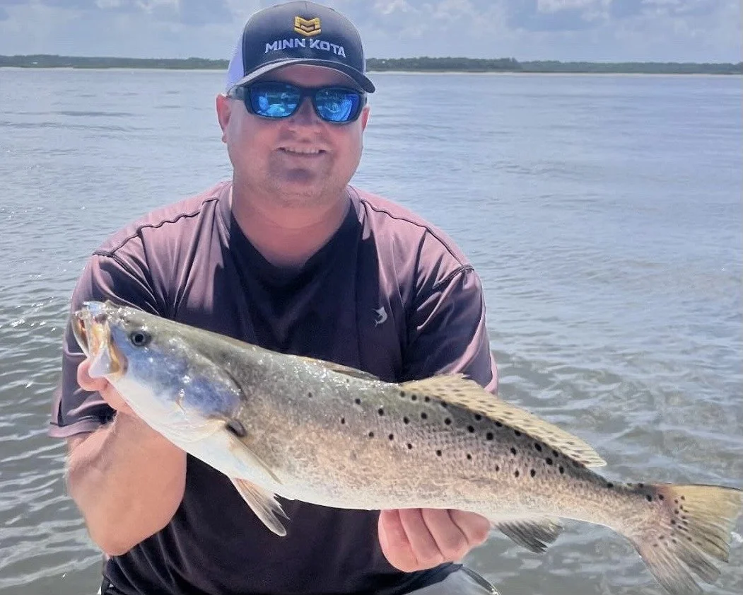 Man in sunglasses and a black cap holding a large fish near a body of water with a cloudy sky.