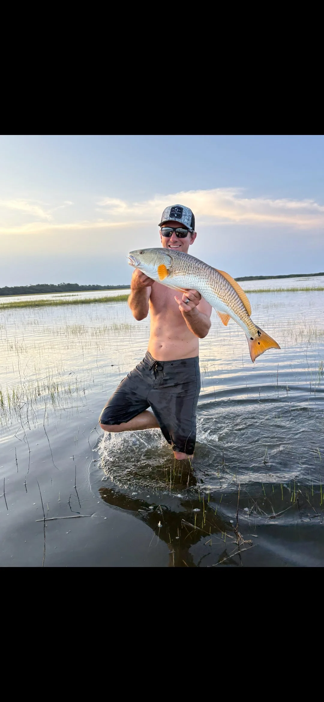 A man standing knee-deep in water, shirtless, wearing sunglasses and a hat, holding a large fish with both hands, smiling at the camera with a partly cloudy sky and marshy landscape in the background.