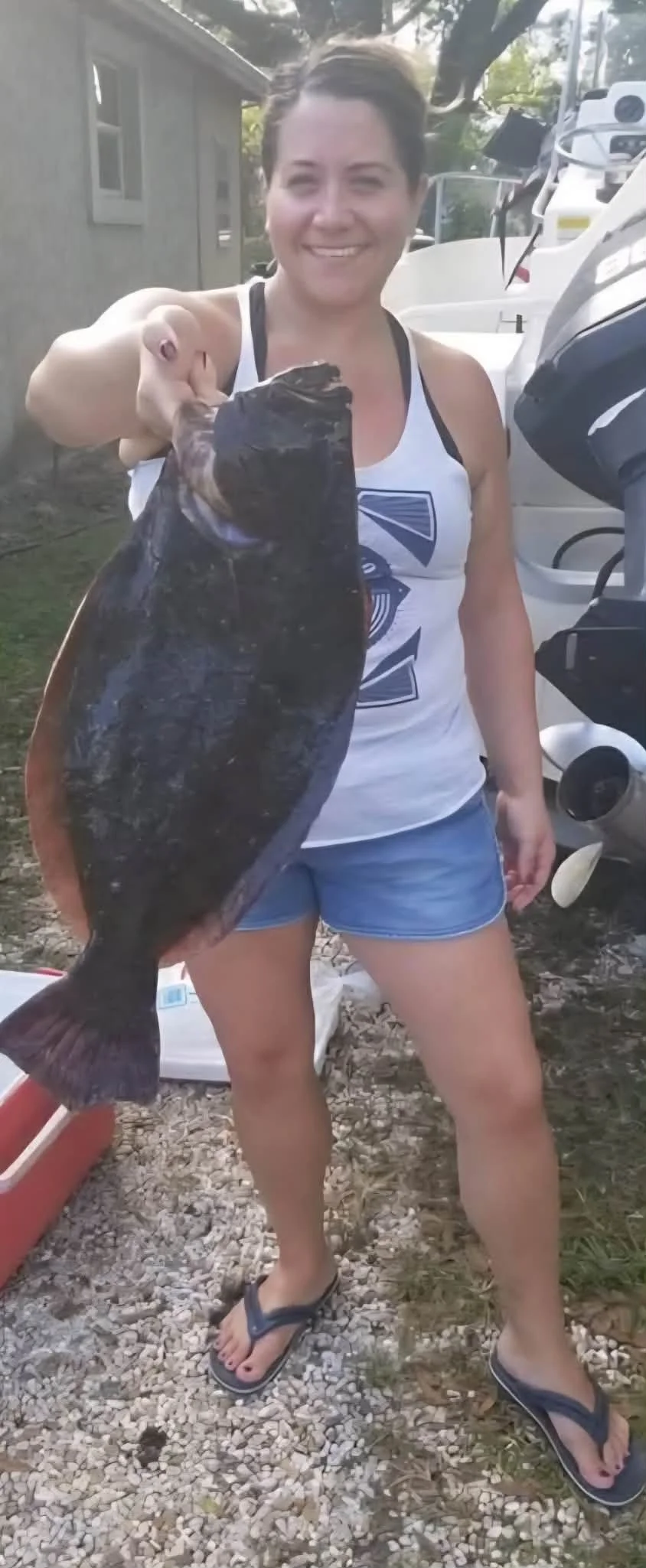 Woman smiling and holding a large fish outdoors near boats and a building.
