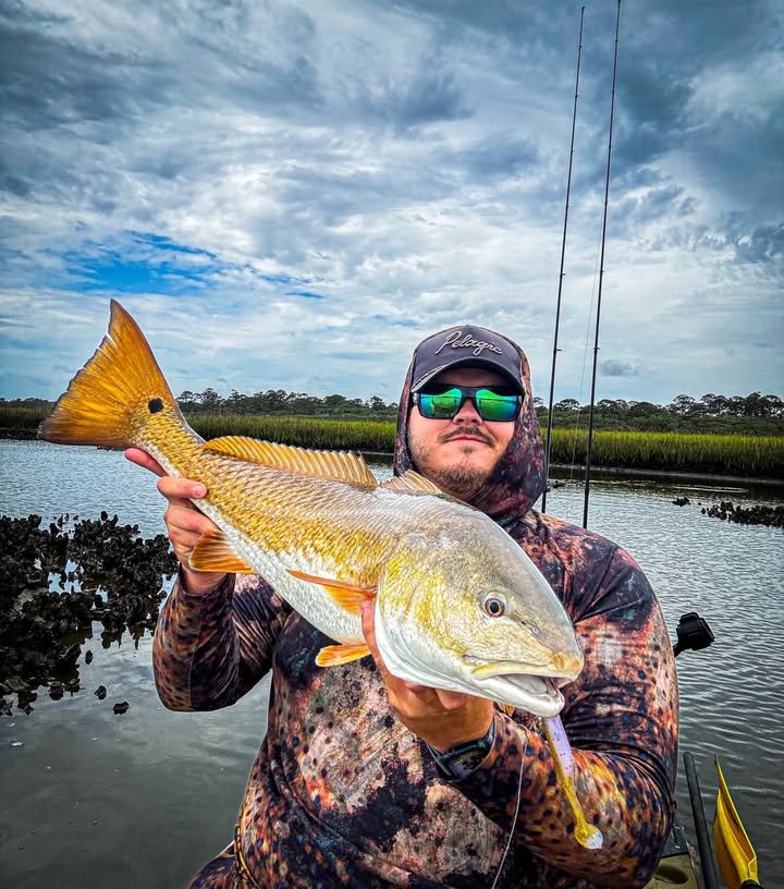 A man in camouflage clothing and sunglasses holding a large fish near a body of water with a cloudy sky.