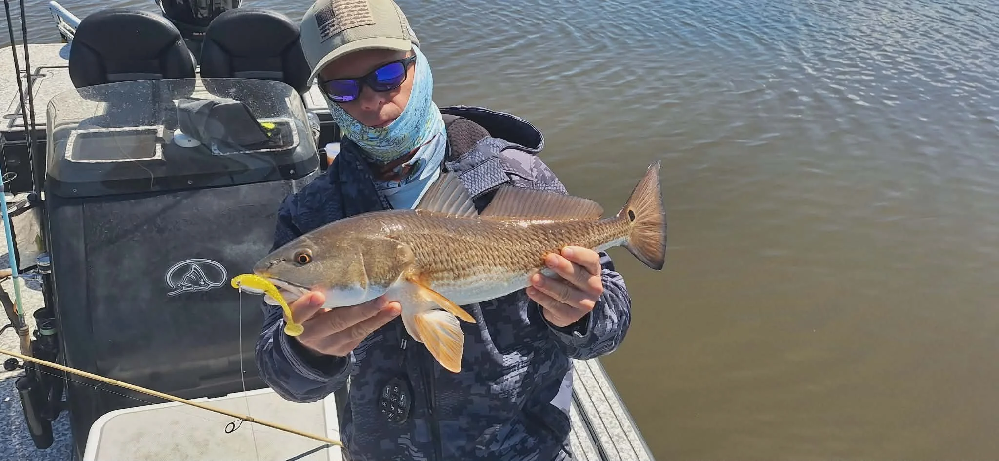 A person holding a large fish on a boat, dressed in sunglasses, a cap, and a camo jacket, with water in the background.