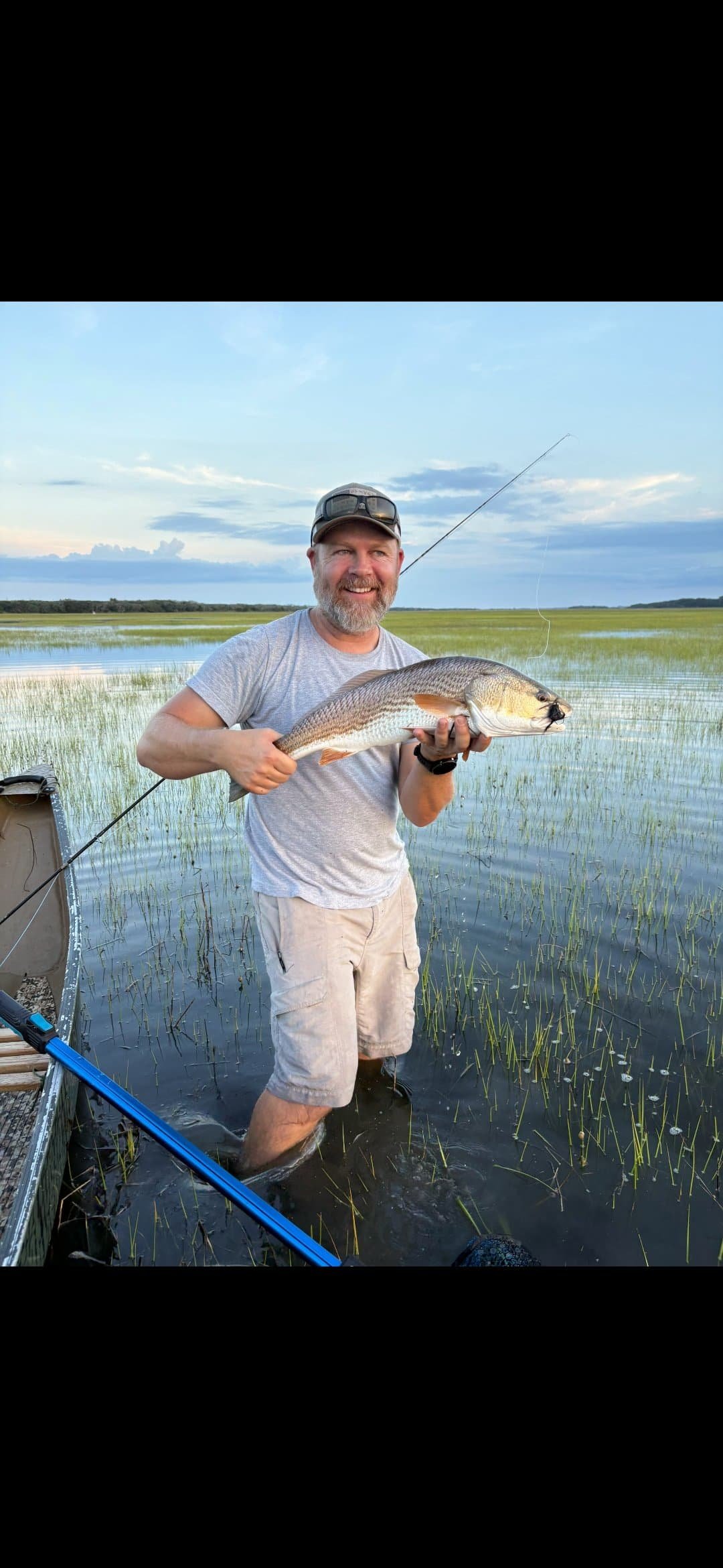 A man with a beard, wearing a gray t-shirt, khaki shorts, and sunglasses on a cap, smiling and holding a large fish while standing in shallow water with grass and a body of water in the background during sunset.