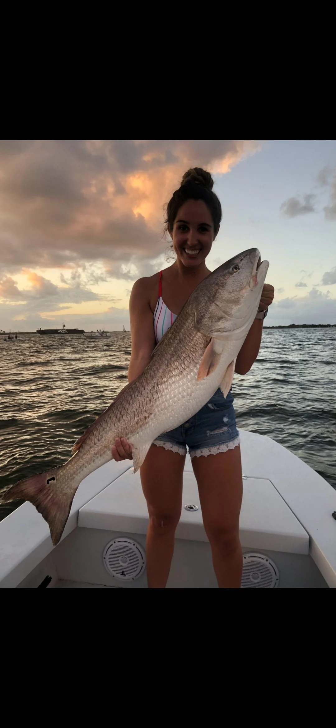 A woman standing on a boat holding a large fish, with a sunset and cloudy sky in the background.