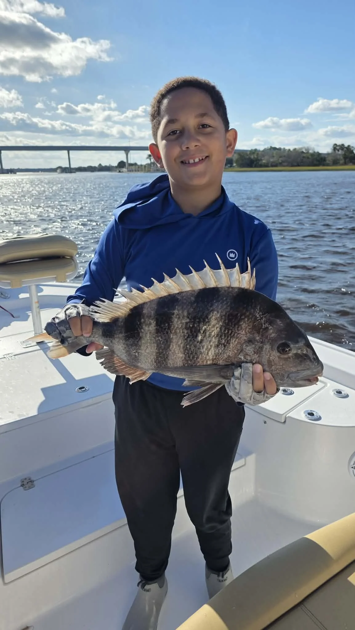 A young boy holding a large fish on a boat, with water, a bridge, and a partly cloudy sky in the background.