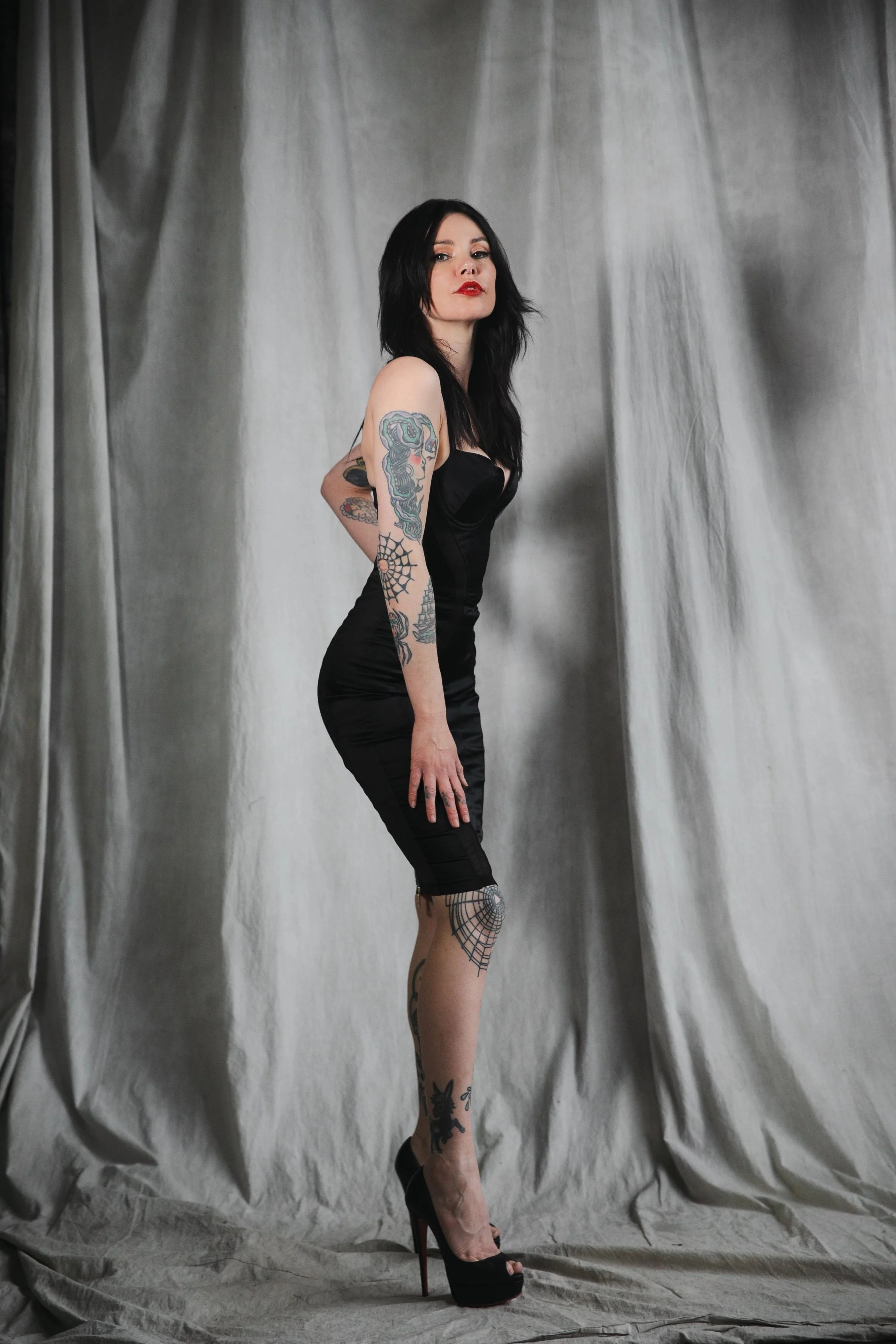 A woman with black hair and tattoos wearing a black dress and high heels, standing in front of a grey curtain backdrop.